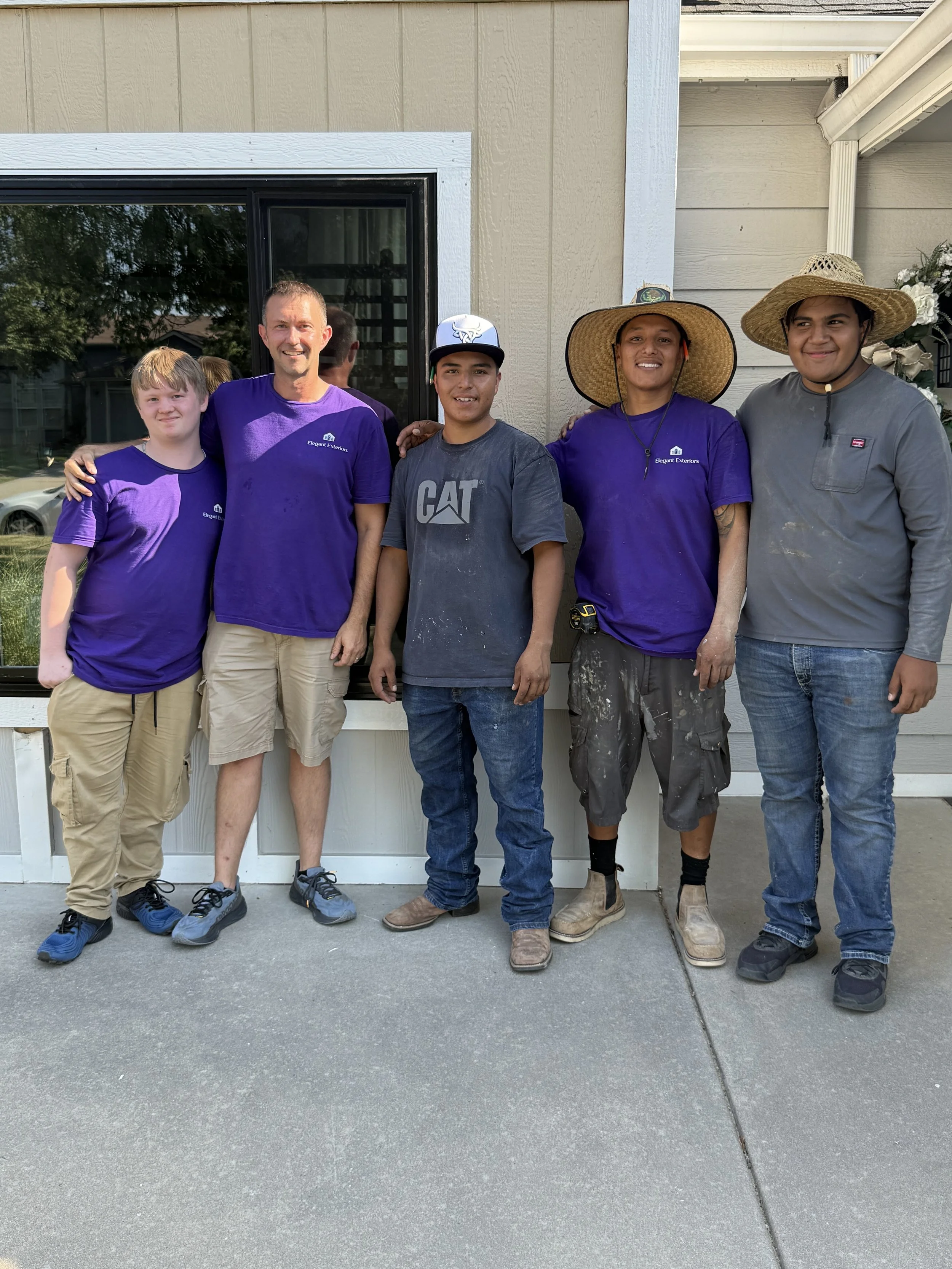 Group of five people standing outdoors in front of a house, smiling for the photo. The group includes three men and two boys, some wearing purple shirts and wide-brimmed hats, with one person in a gray shirt with dirt marks.