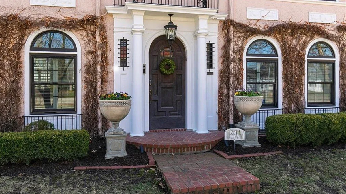 Exterior of a house with arched windows, a black front door with a green wreath, white columns, two large planters with flowers, and neatly trimmed bushes.