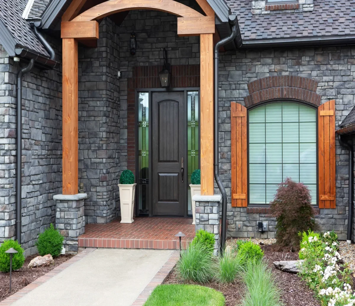 Front entryway of a house with a black door, stone exterior, wooden shutter accents, and a small landscaped garden with plants and shrubs.