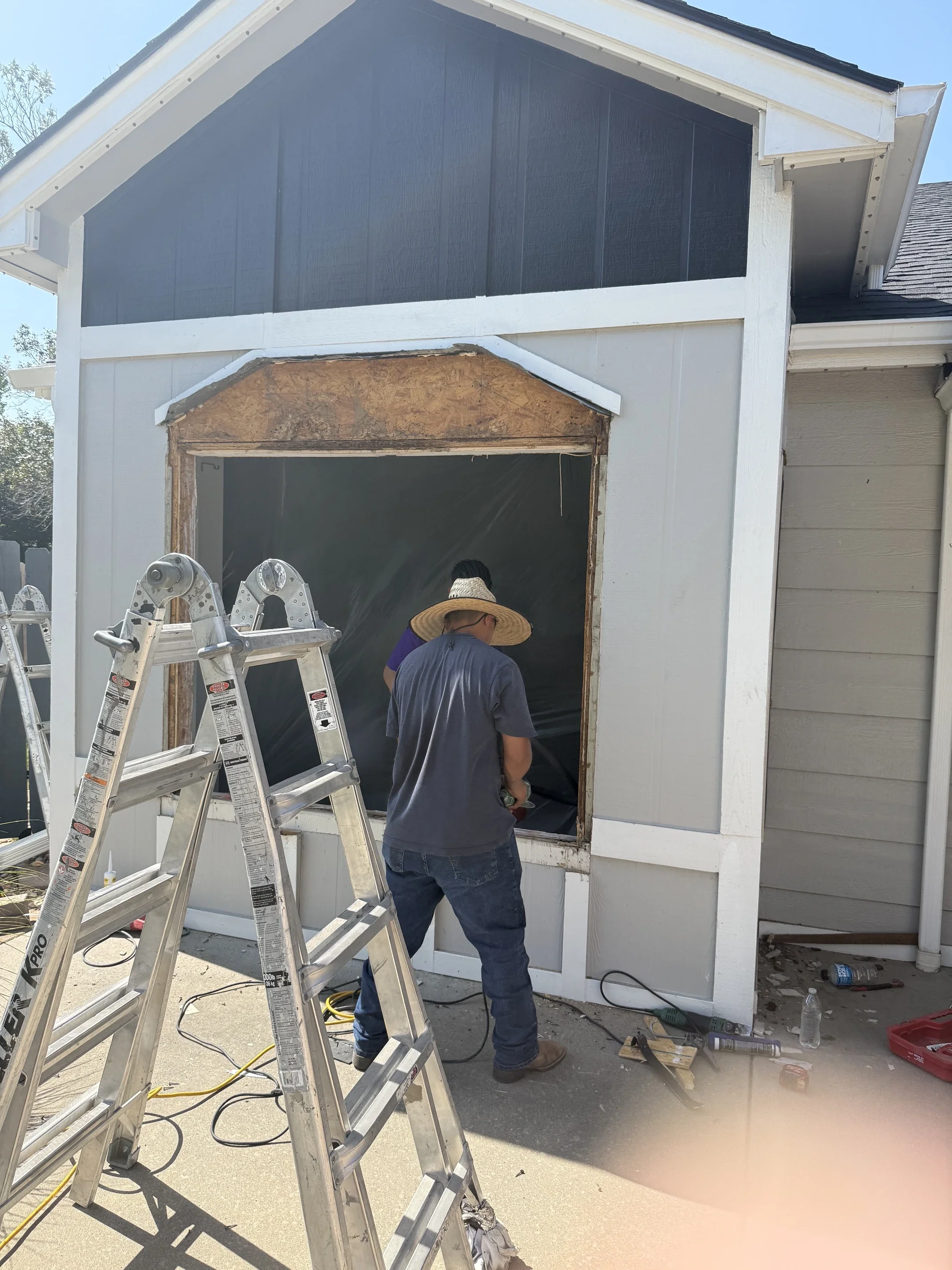 Two workers are installing a window in a house, with ladders nearby and construction tools on the ground.