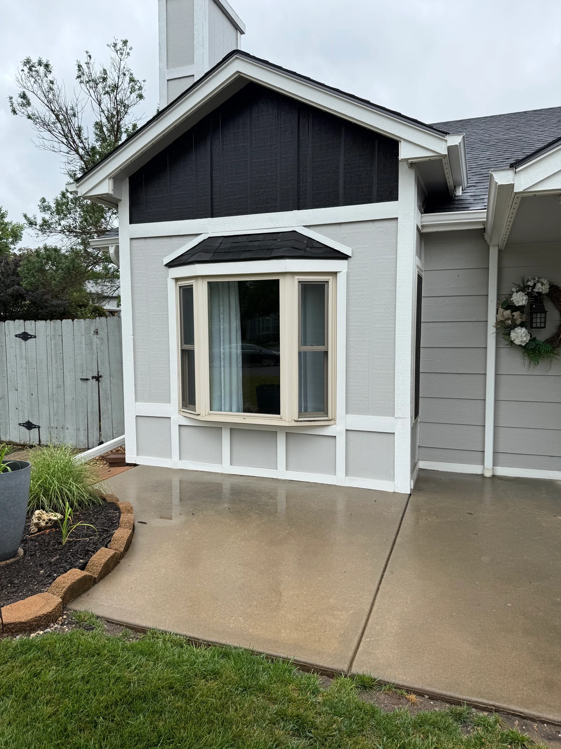 View of the side of a house showing a bay window and a black-paneled gable with white trim, with a concrete patio in front, a small garden bed, and a wooden fence to the left.