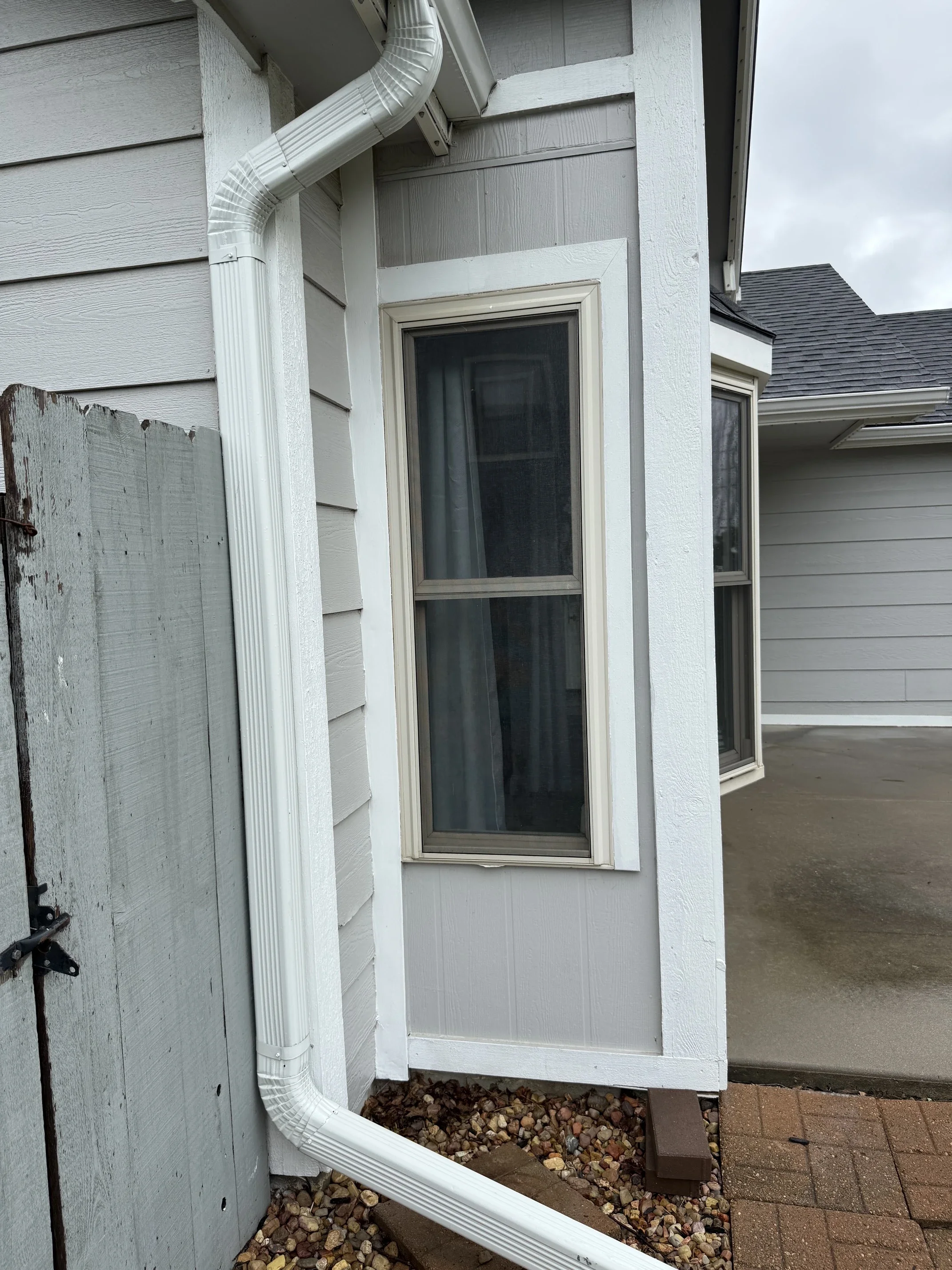 Outside corner of a house showing a downspout connected to an outer wall with a window, gravel on the ground, and part of a concrete walkway and brick pavers.