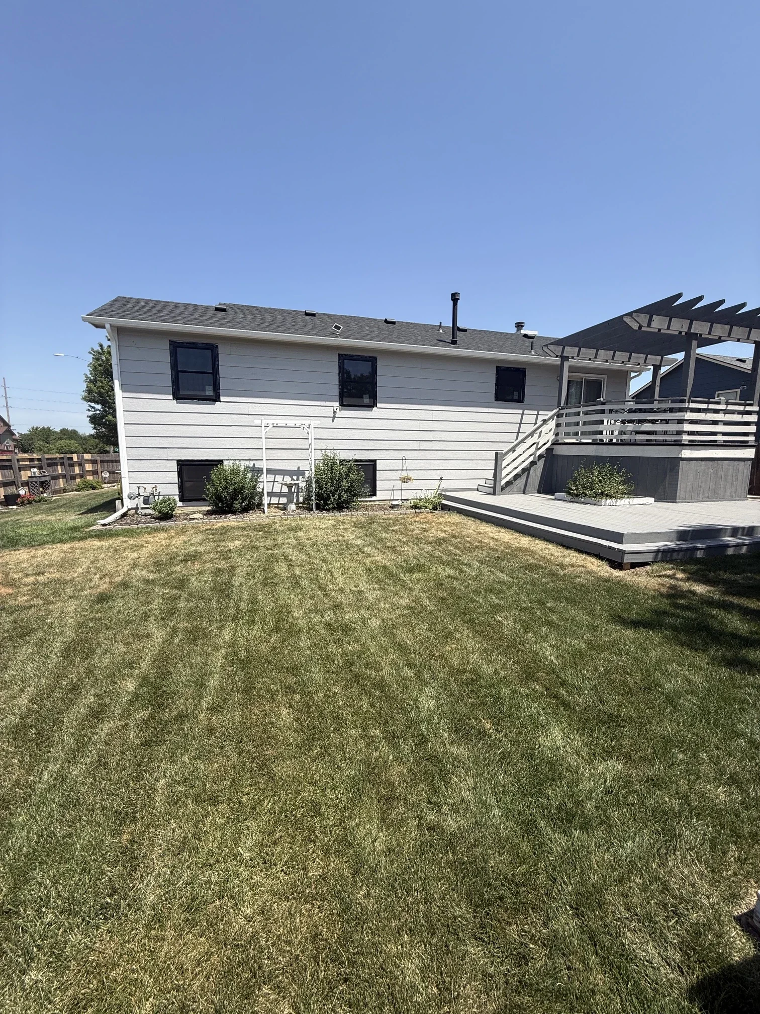 A backyard with a well-maintained lawn, a white two-story house with black windows, a wooden deck with stairs, and a clear blue sky.
