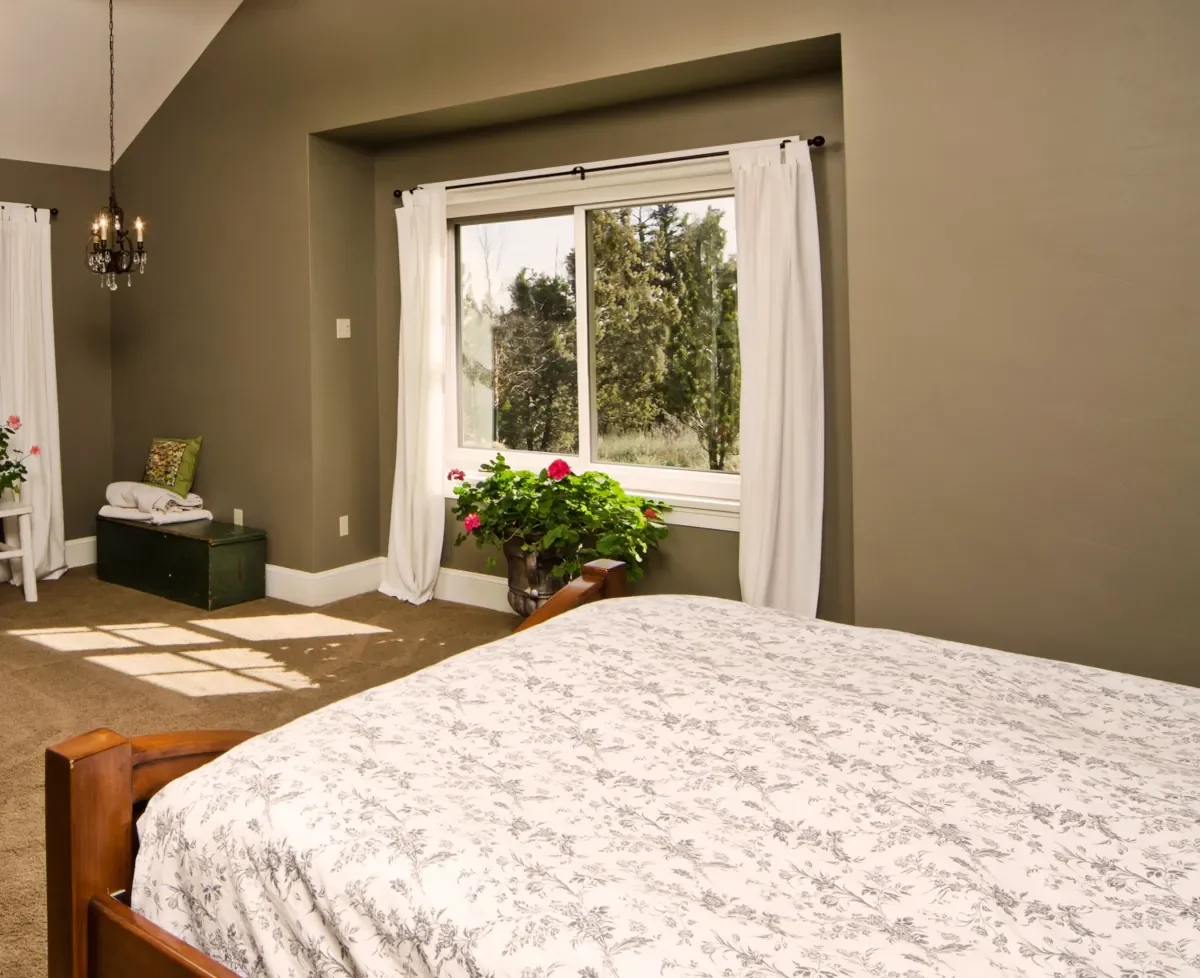 A bedroom with a bed in the foreground, white floral bedding, a large window with white curtains, a potted plant with pink flowers, and a view of trees outside.
