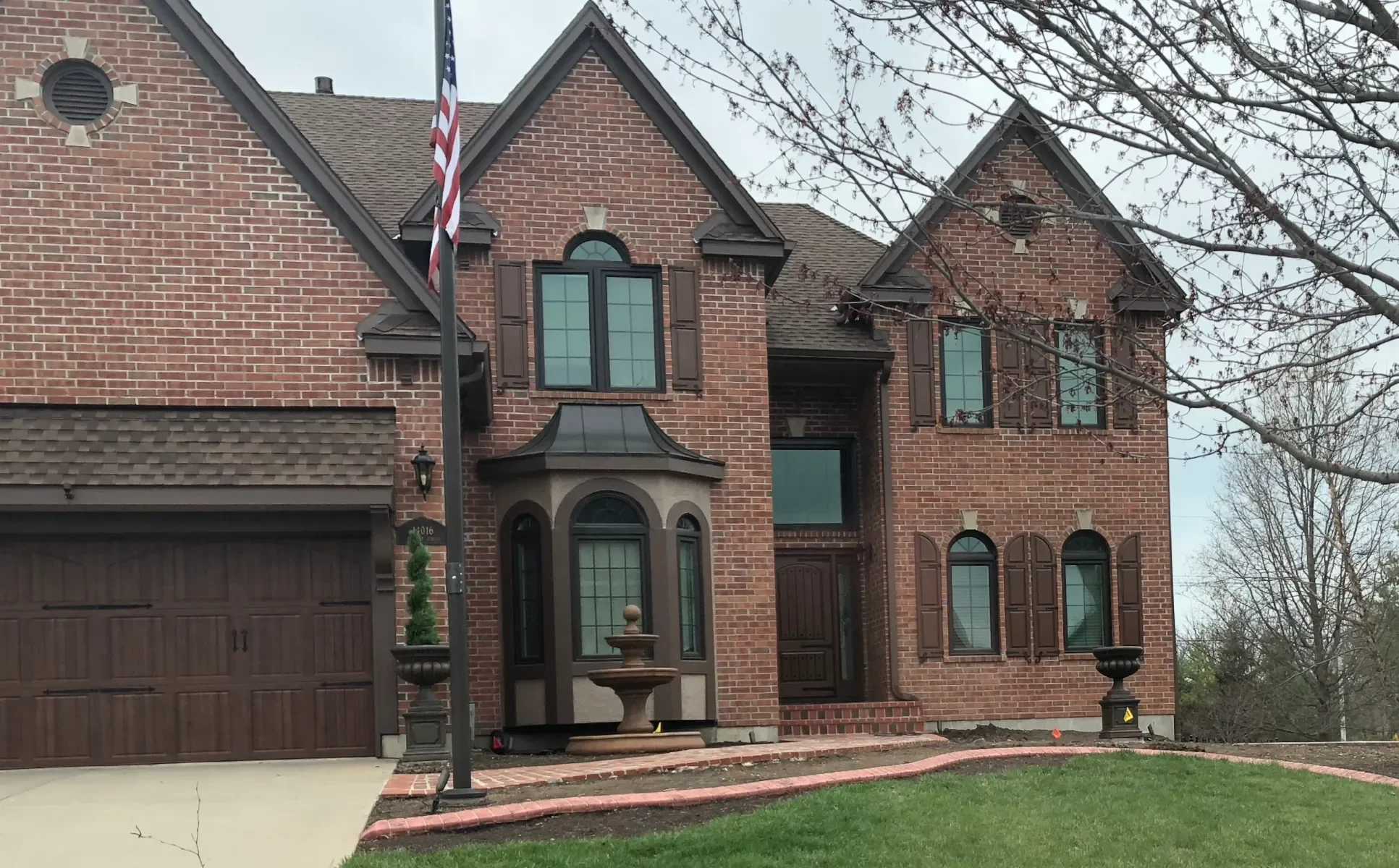 A large brick house with a dark brown garage door, multiple windows with brown shutters, and a front porch with a decorative fountain and small potted plants. An American flag is on a pole near the sidewalk, with leafless trees and overcast sky in the background.