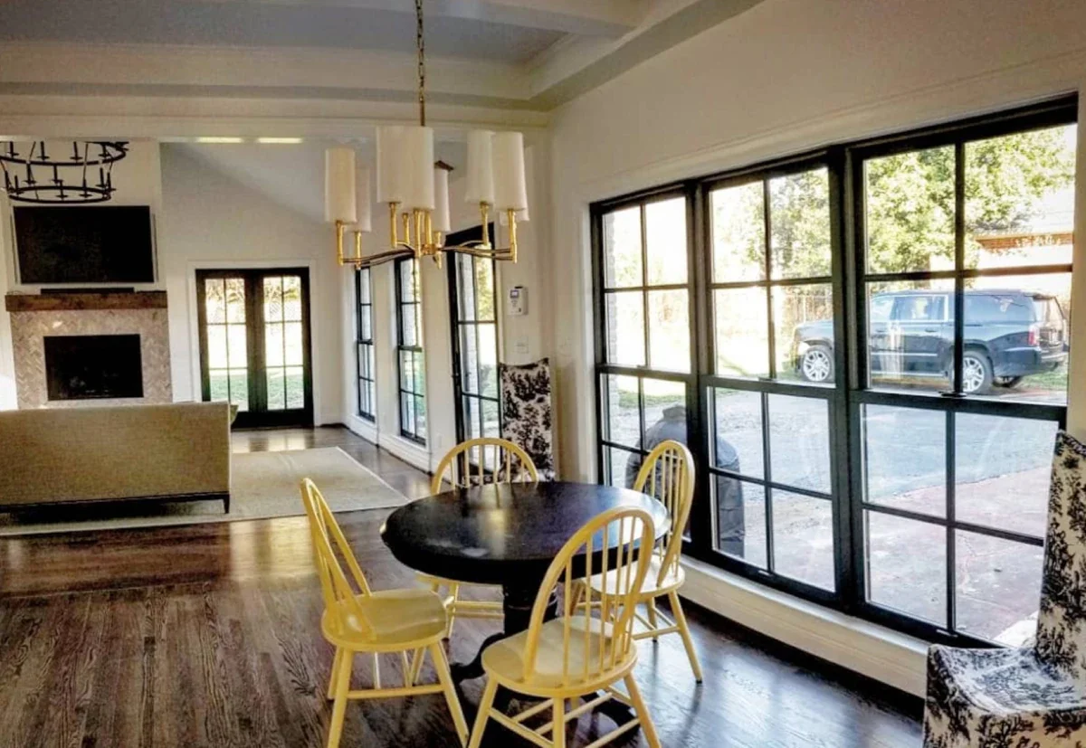 Bright living room with large windows, a dark round dining table with four yellow chairs, floral patterned chairs, a beige sofa, a fireplace, and a wall-mounted TV, with hardwood floors and modern decor.