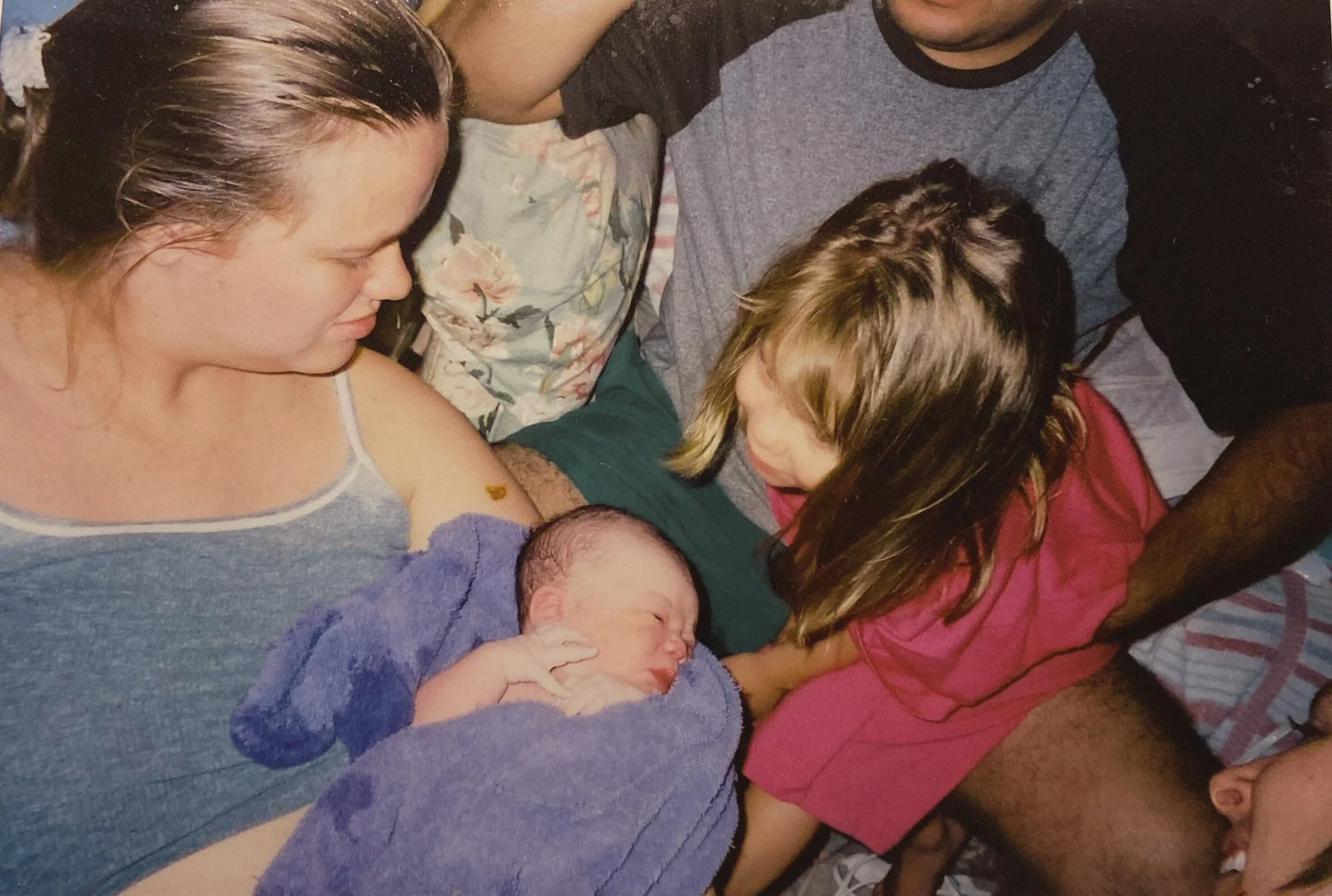 A woman holding a newborn baby wrapped in a purple blanket, with a young girl and a man nearby, all gathered around the baby in an intimate setting.