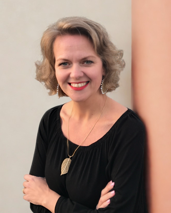 A woman with shoulder-length curly blonde hair smiling, wearing a black top, silver earrings, a gold leaf pendant necklace, and pink nail polish, standing against a light-colored wall.