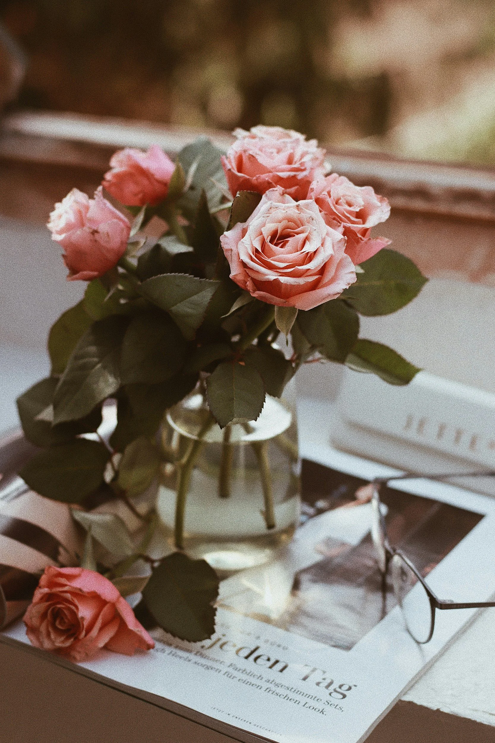 A bouquet of pink roses in a small glass vase placed on top of magazines and papers on a table. A single rose is lying on the surface next to the bouquet, and glasses are resting on one of the magazines.
