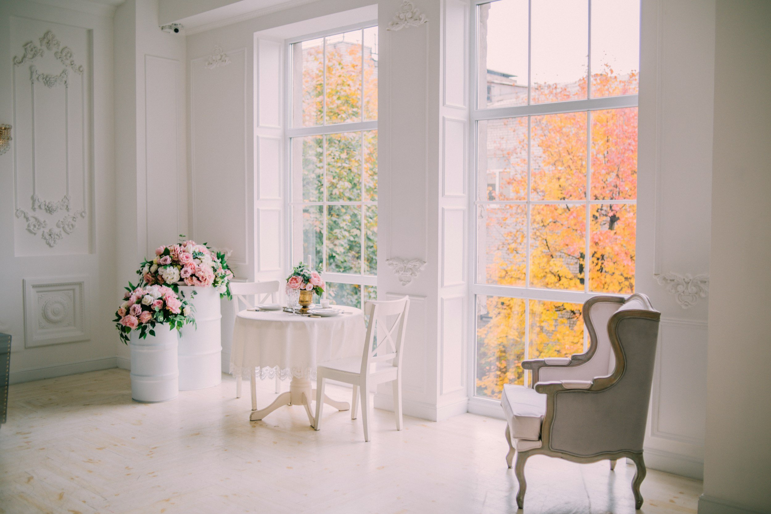 Bright white room with large windows, pink floral arrangements, a small dining table with chairs, and an elegant armchair with a view of fall foliage outside.