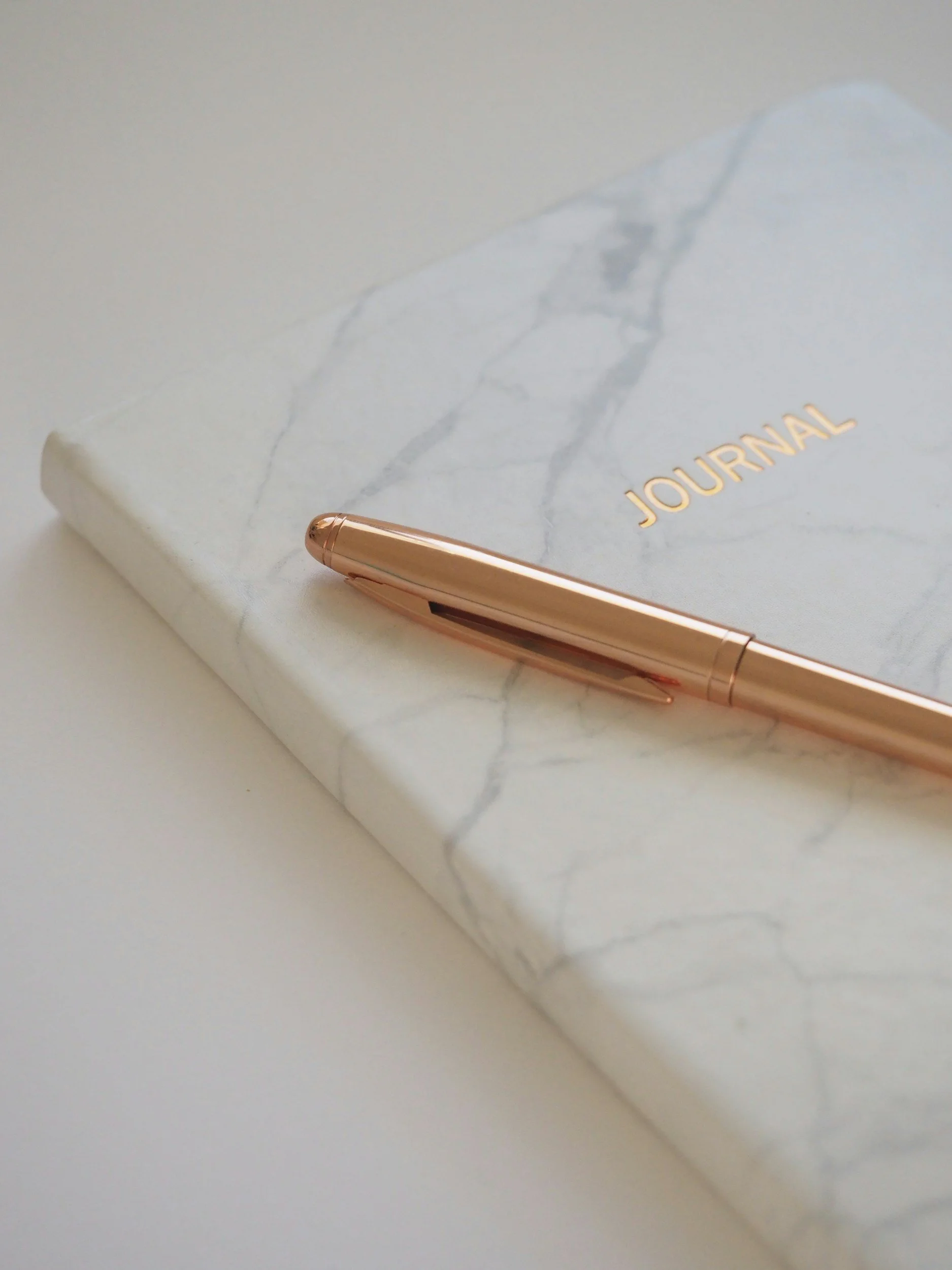 A marble-covered journal with the word 'JOURNAL' in gold on the cover, and a rose gold pen resting on it.
