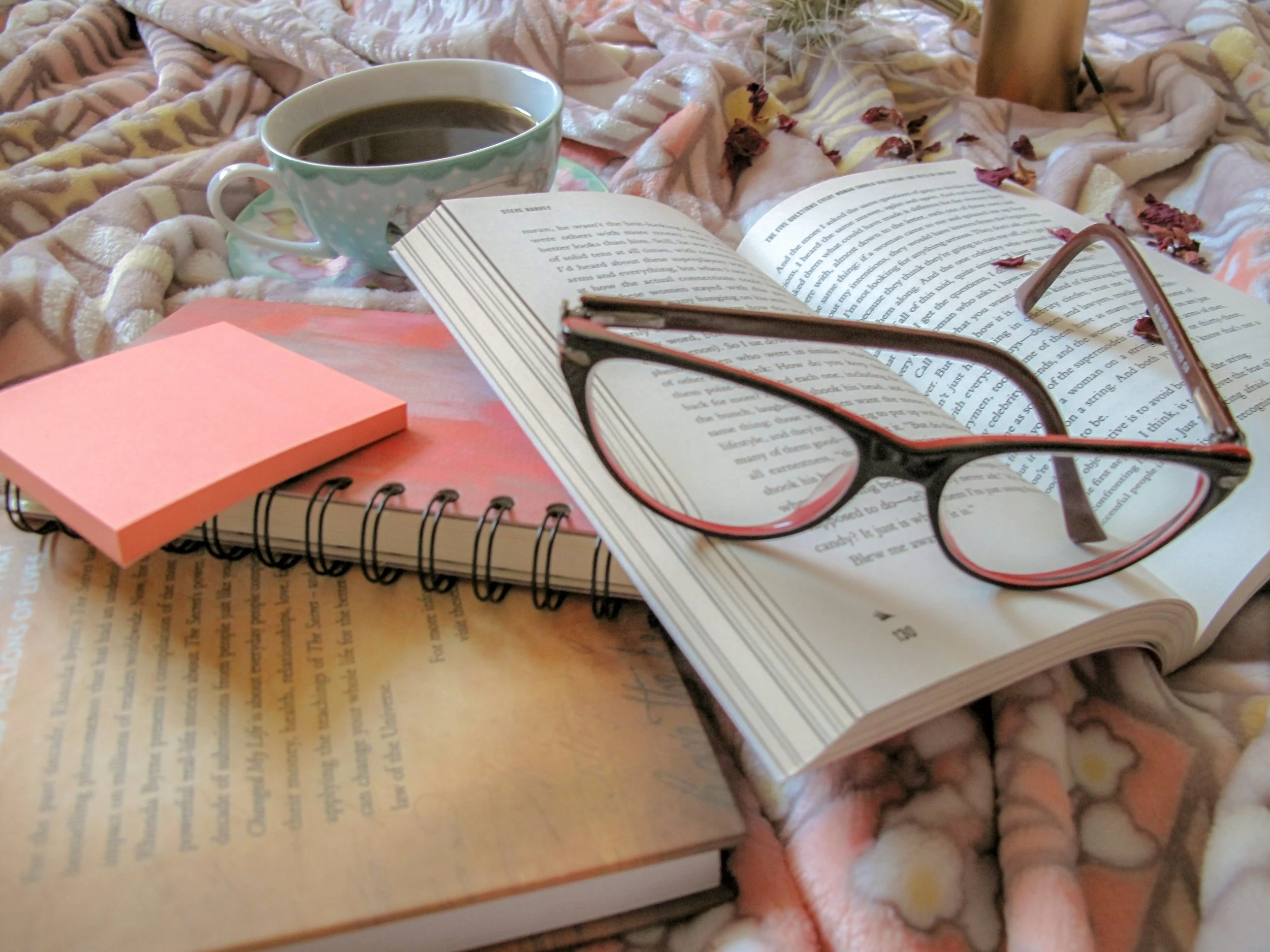 Open book with reading glasses on top, a smartphone, a notebook, a cup of coffee, and pink sticky notes on a bed covered with a floral blanket.
