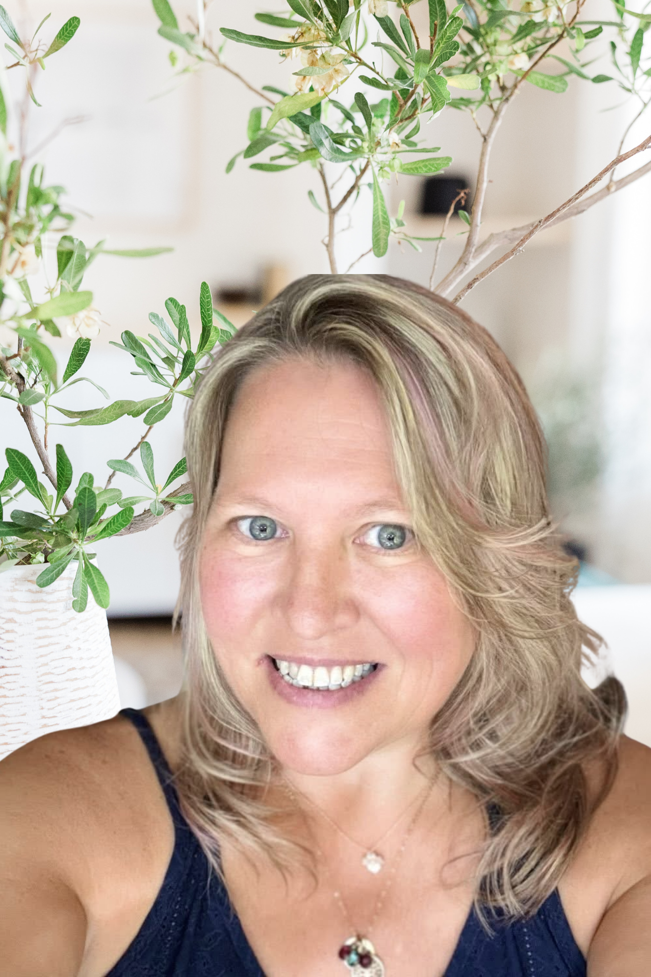 A smiling woman taking a selfie in front of a potted plant with green leaves and white flowers.