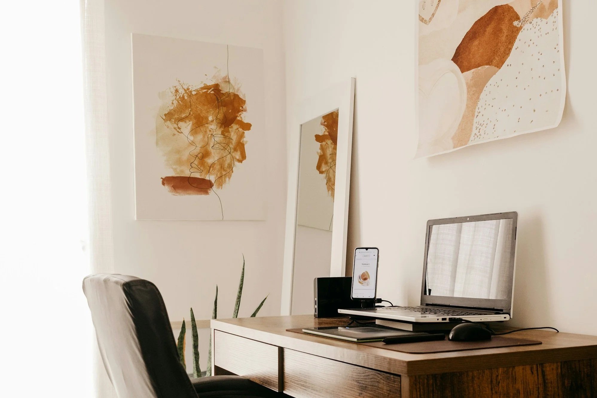 Home office with a wooden desk, a laptop, an external monitor, a mouse, and a smartphone. Art prints and a mirror are on the white wall behind the desk. There is a black office chair and a potted plant on the left side.