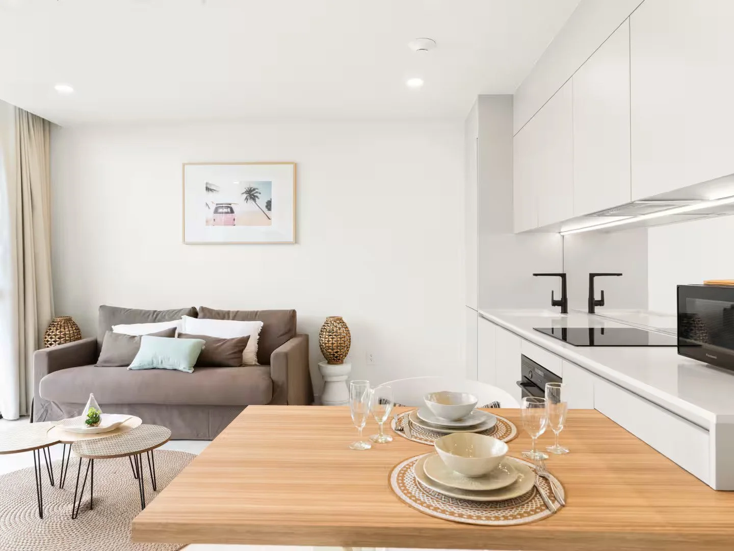 Open-plan living room and kitchen with beige sofa, round coffee tables, dining table with bowls and glasses, white cabinets, black stovetop and microwave, wall art, and curtains.