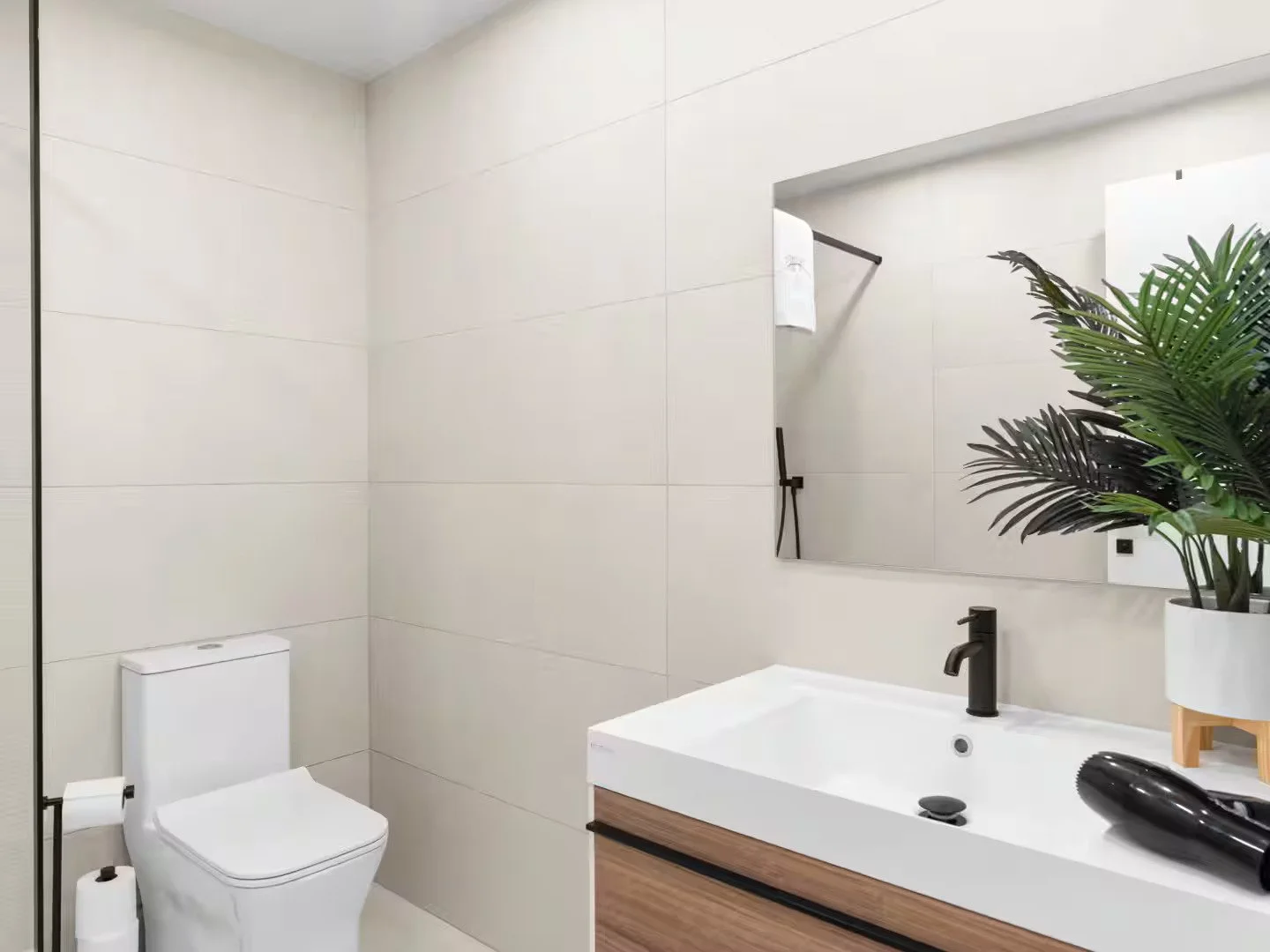 Modern bathroom with a white toilet, a large mirror, a rectangular sink with a black faucet, a black hairdryer, a green potted plant, and cream-colored tiled walls.