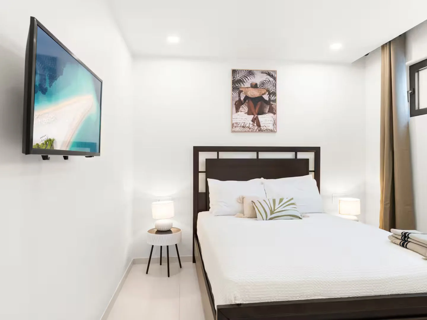 Minimalist bedroom with white walls, a bed with white bedding, a black headboard, two bedside lamps, a picture of a person in a hat above the bed, a flat-screen TV on the wall, and a window with beige curtains.