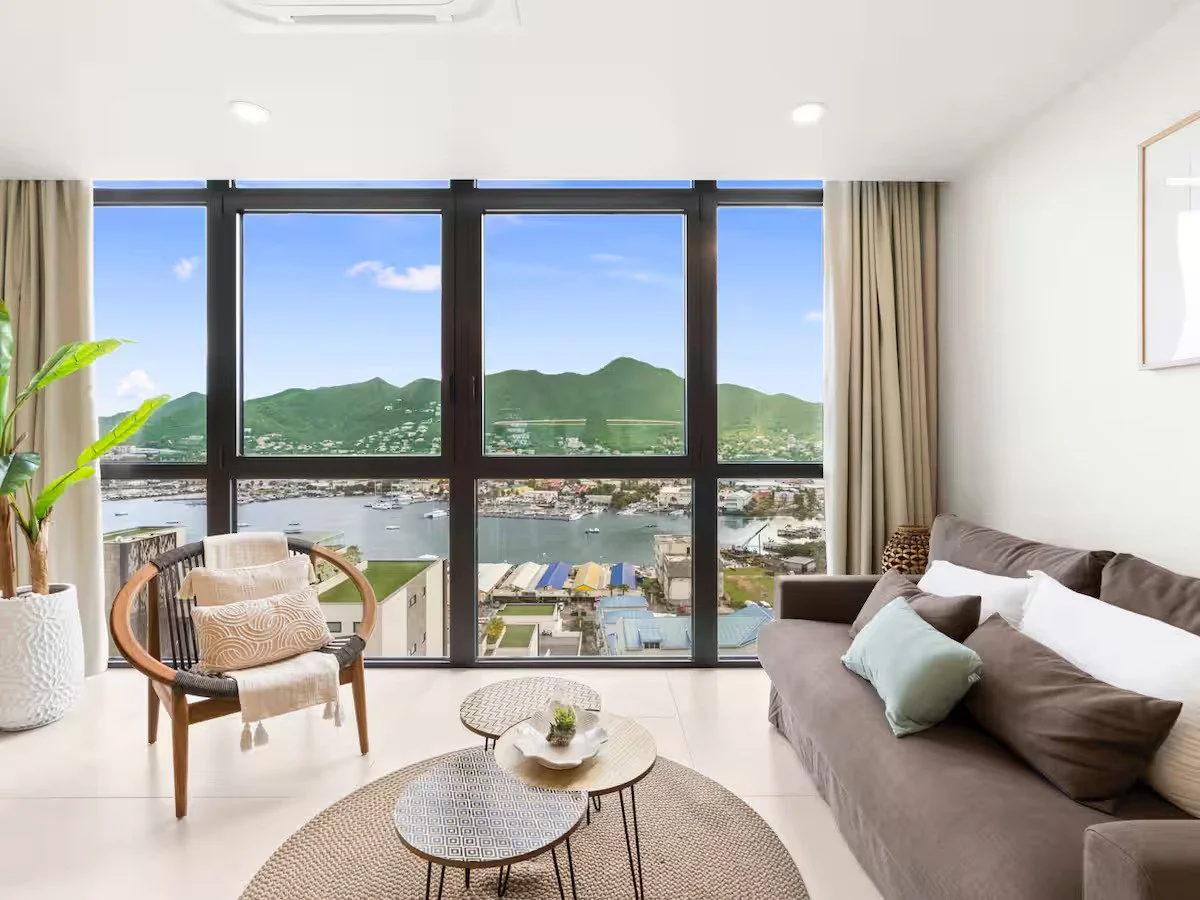 Living room with large window overlooking a river, mountains, and a cityscape, furnished with a gray sofa, a wooden chair with a cushion, and small coffee tables, decorated with plants and curtains.