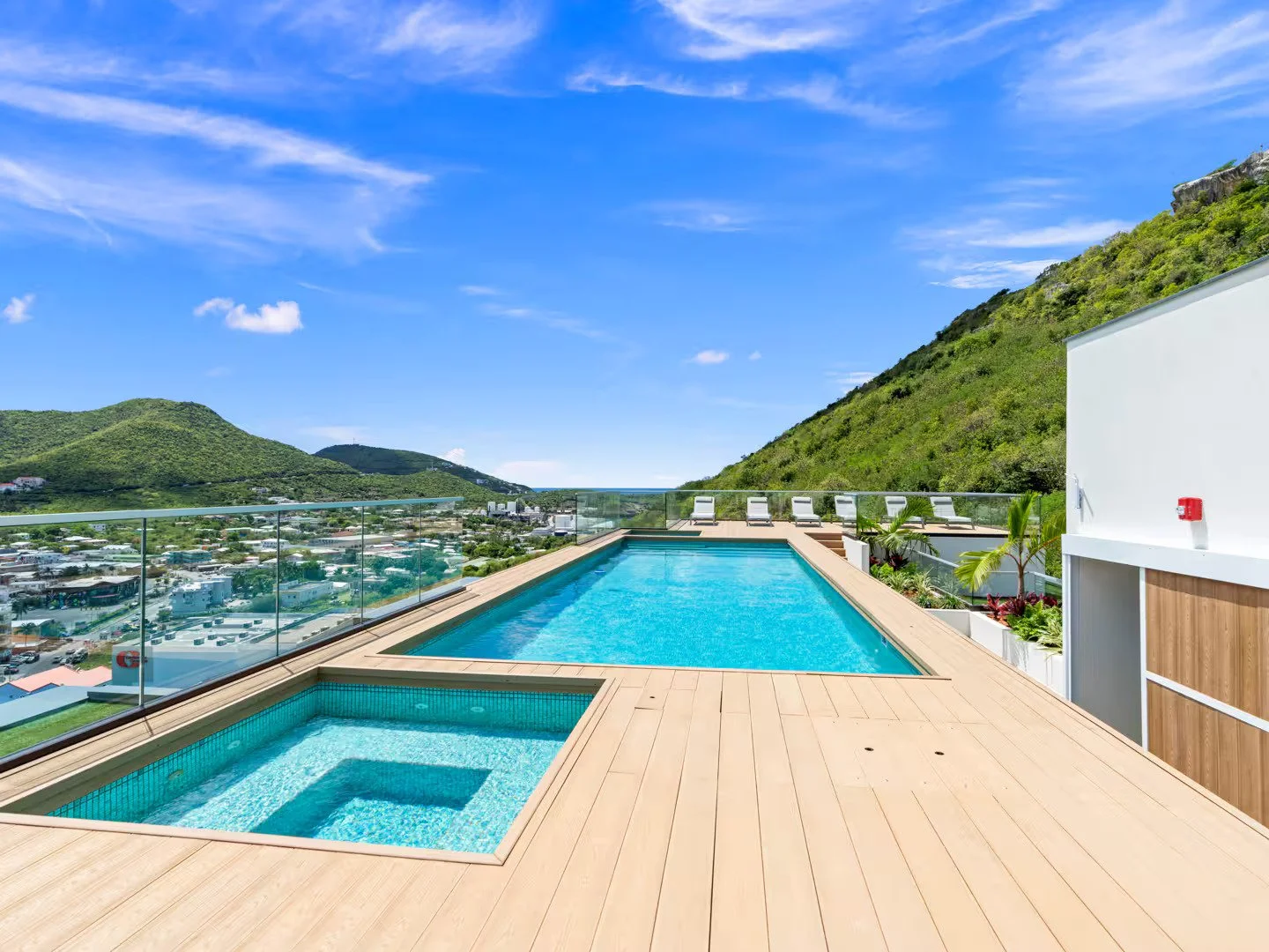 Rooftop infinity pool overlooking green hills and a town under a blue sky with clouds.
