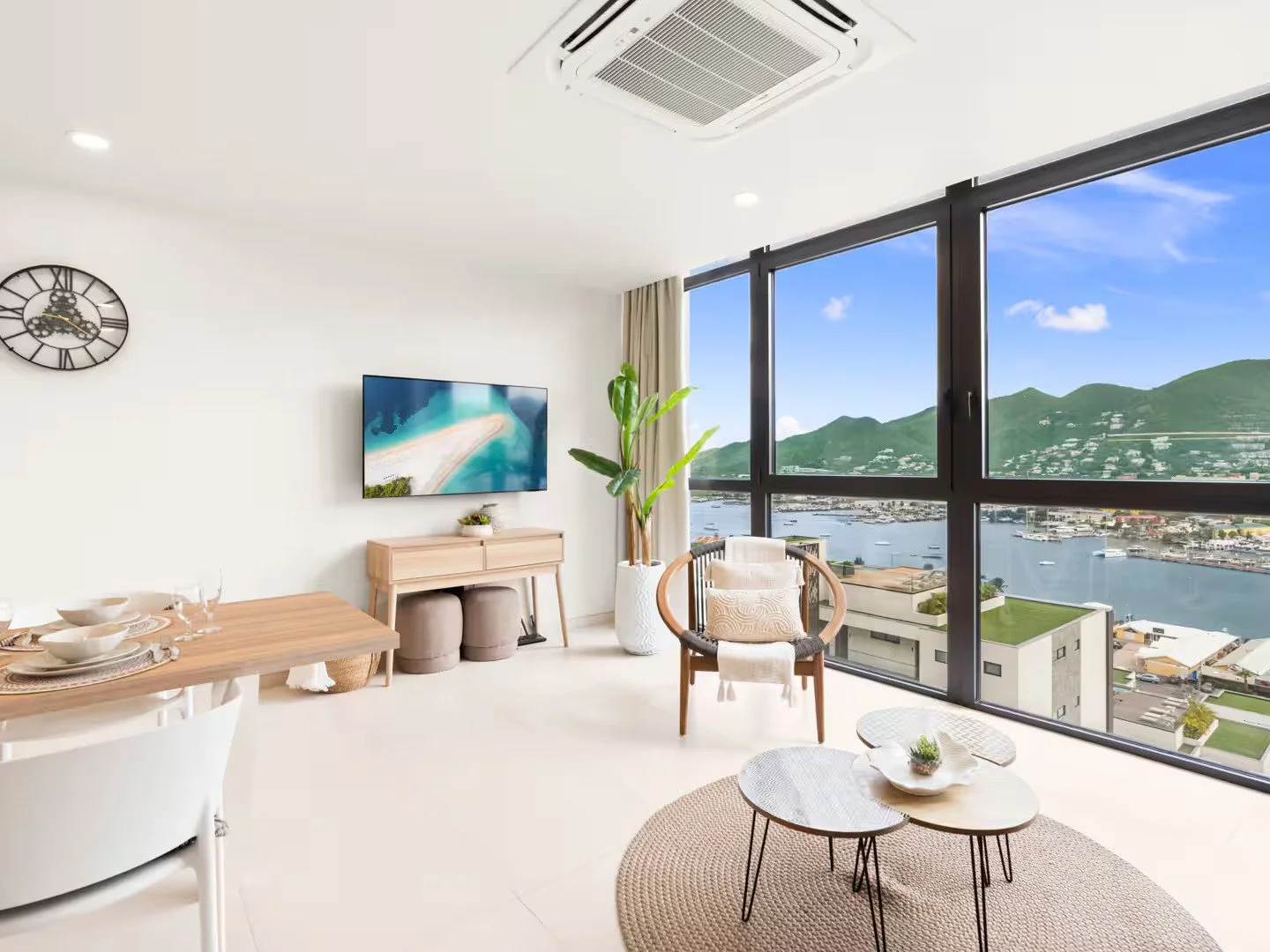 Modern living room with large floor-to-ceiling windows offering a view of the city harbor and green mountains, featuring a wall clock, flat-screen TV, potted plant, and minimalist furniture.