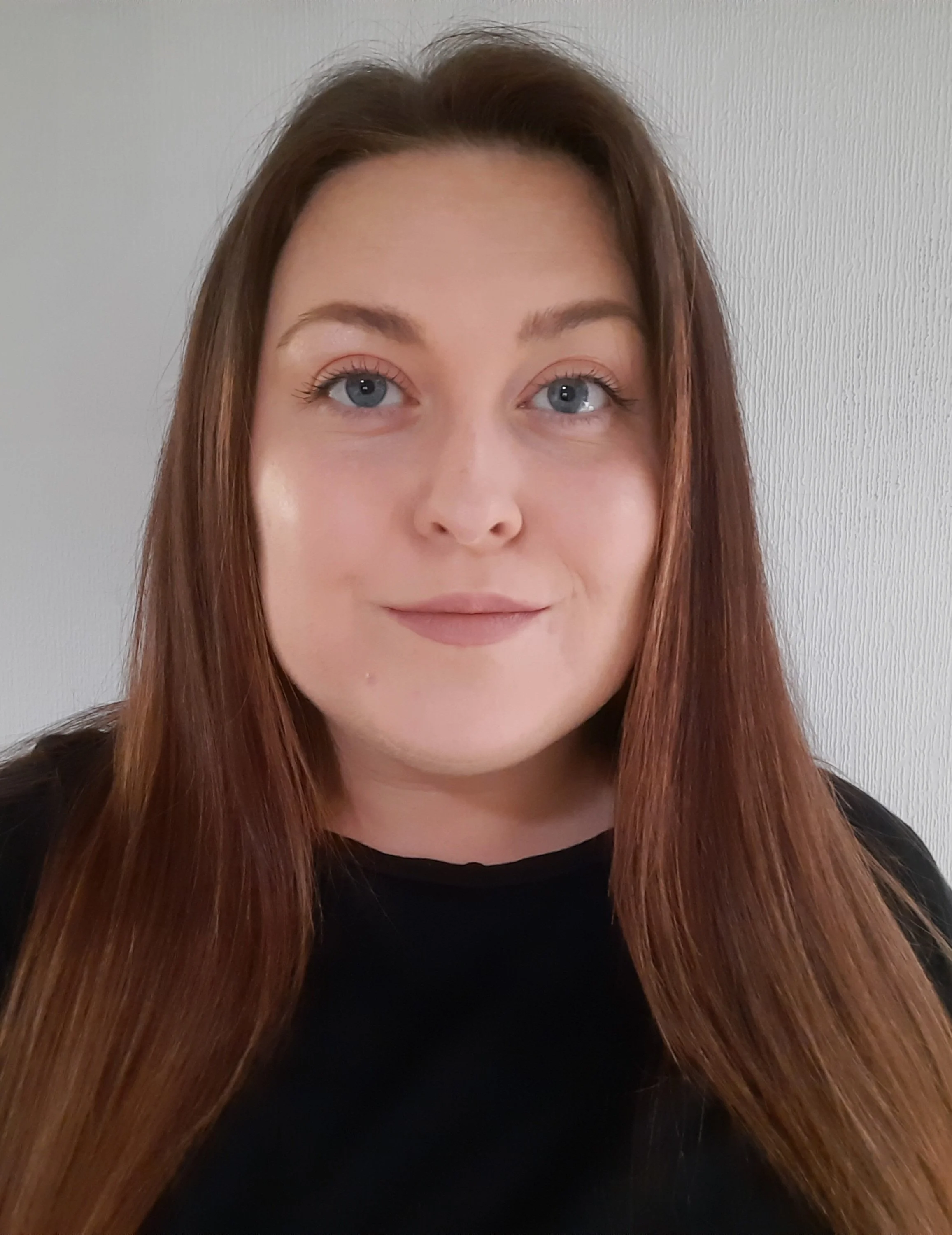 Close-up of a woman with long, straight red hair and blue eyes, wearing a black top, standing against a plain white wall.