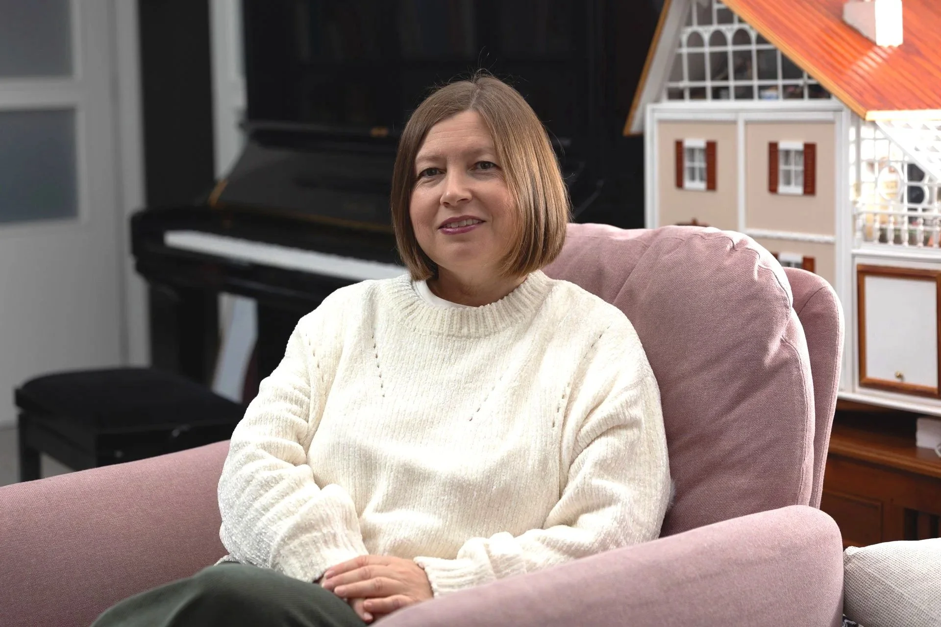 Mª Ángeles sentada en un sillón rosa, con un piano negro y un mobiliario decorativo en el fondo.