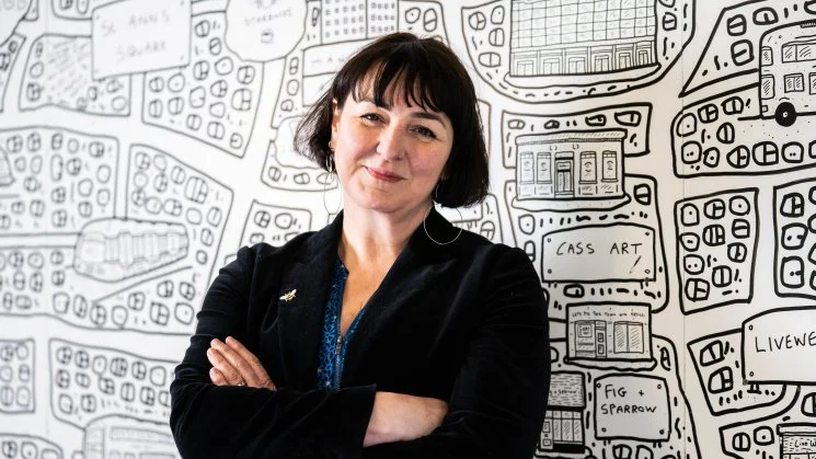 A woman with short dark hair, wearing a black blazer and blue shirt, standing with her arms crossed in front of a detailed black and white city map wall illustration.