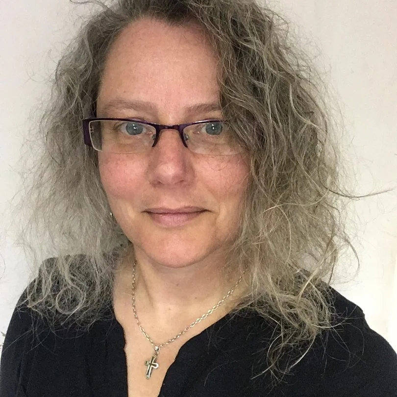 A woman with curly gray hair, wearing glasses, a black top, and a cross necklace, smiling softly against a plain background.
