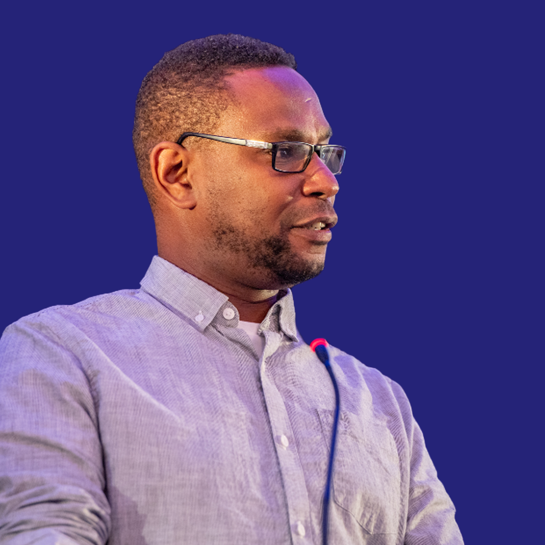 Side profile of a man with glasses and a short fade haircut, wearing a light-colored button-up shirt, with a headset microphone, against a dark blue background.