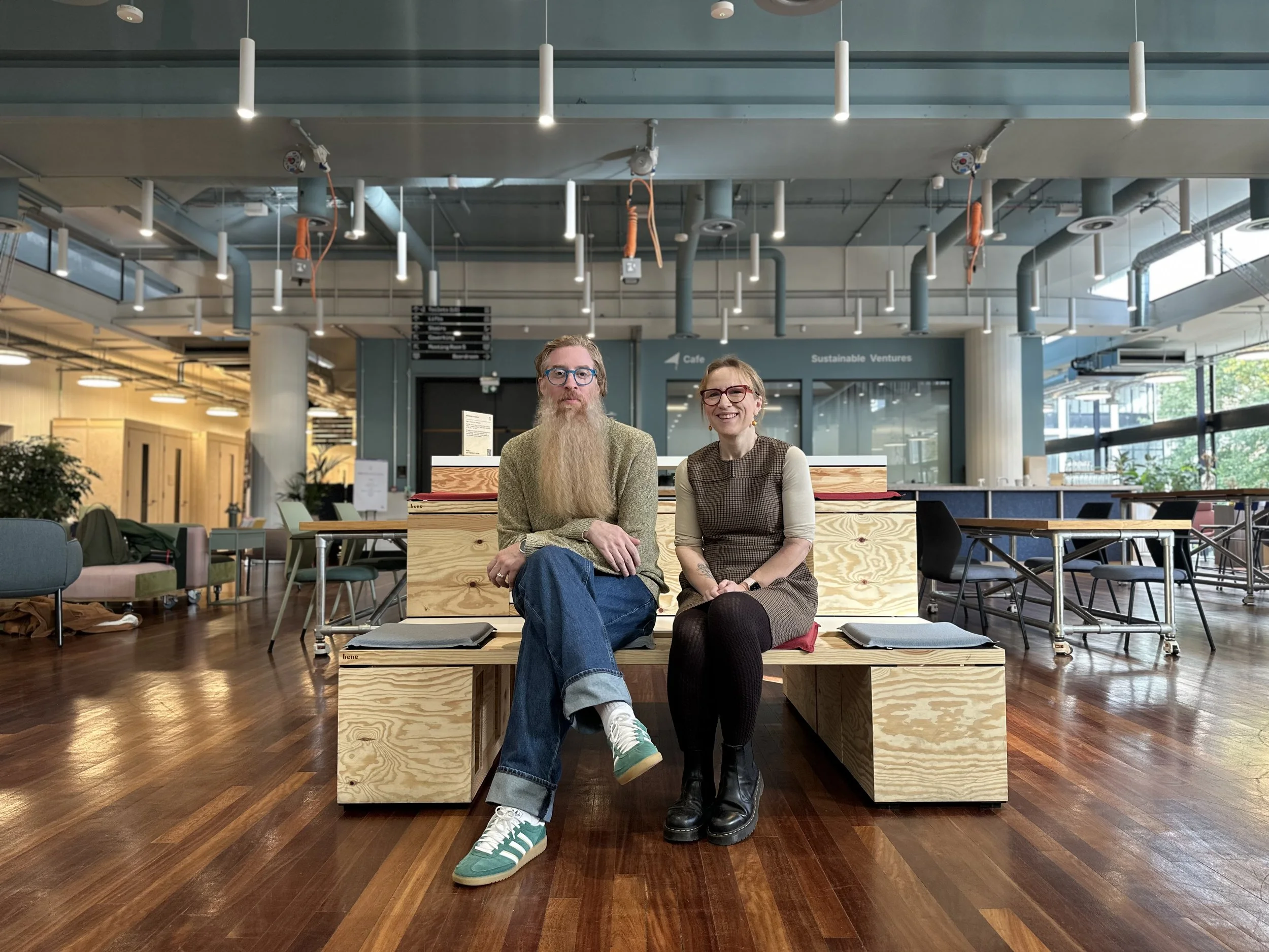 A man with a long beard and glasses sits on a wooden bench next to a woman with glasses and a dress, smiling in a modern, open indoor space with wooden floors, high ceilings, and large windows.