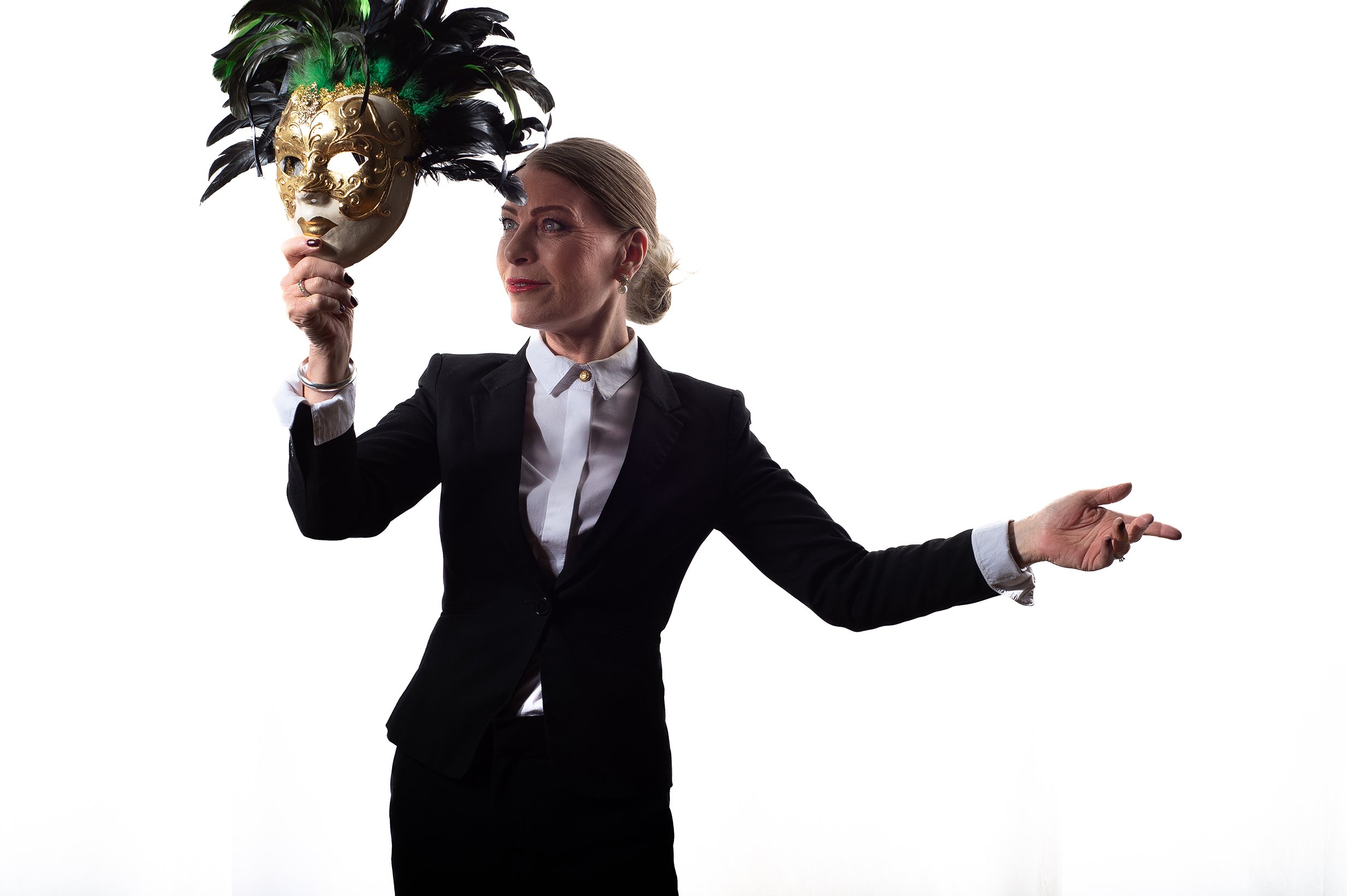 En kvinna i svart kavaj och vit skjorta håller ett maskerat vackert maskeradmask i handen, står mot en vit bakgrund.