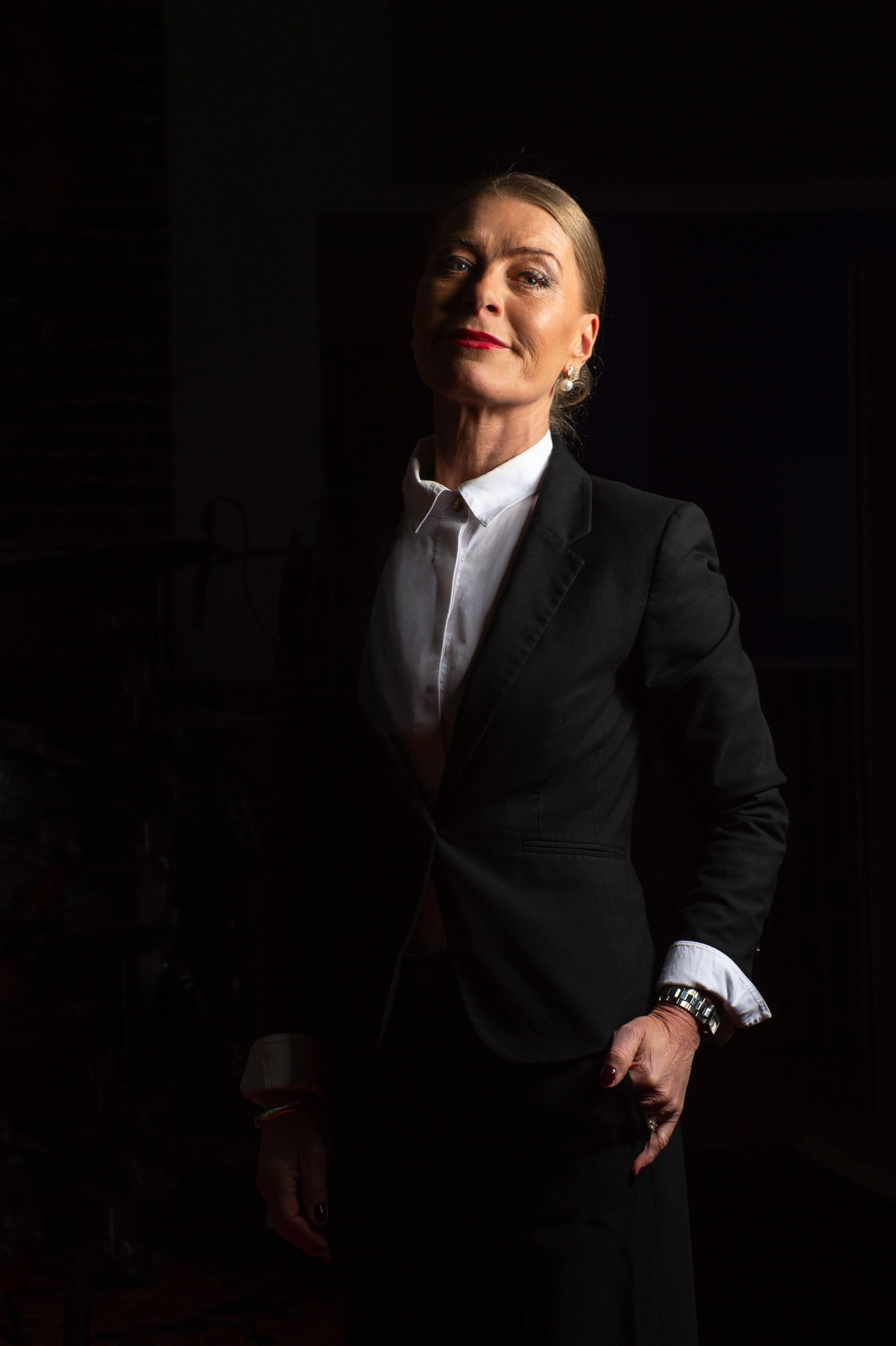 En kvinna i svart kostym och vit skjorta står i ett mörkt rum, belysning fokuserar på hennes ansikte och skapar en dramatisk effekt.