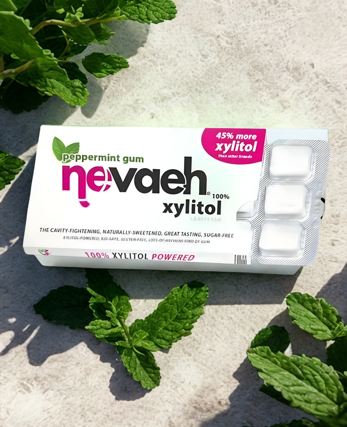 A box of Nevad xylitol peppermint gum on a light surface, surrounded by fresh mint leaves.