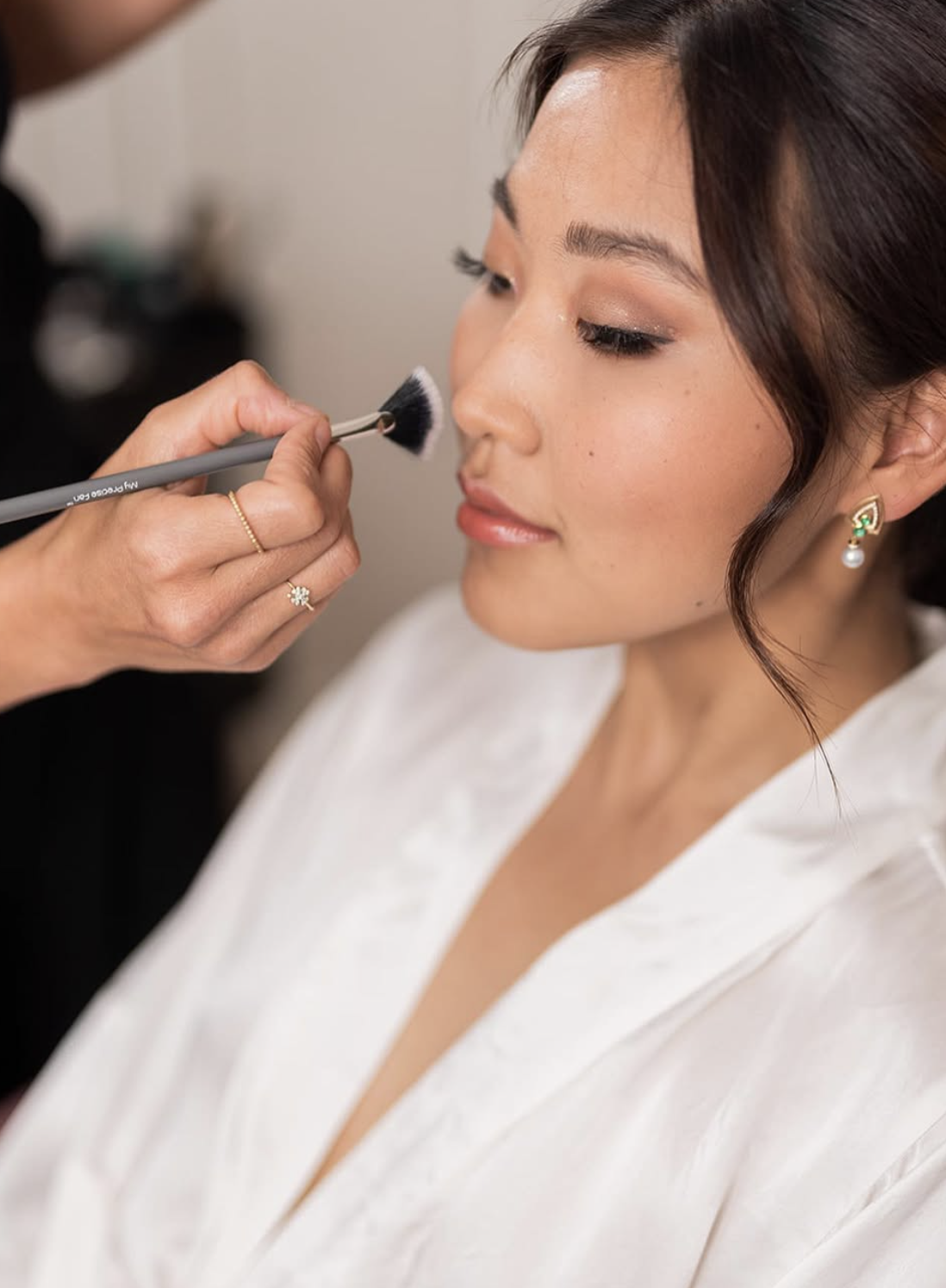 A woman with perfect makeup and jewelry getting her face touched up with a makeup brush.