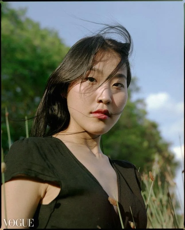A woman standing outdoors in a field with green trees and plants, wearing a black top, with wind blowing her hair, during the daytime.