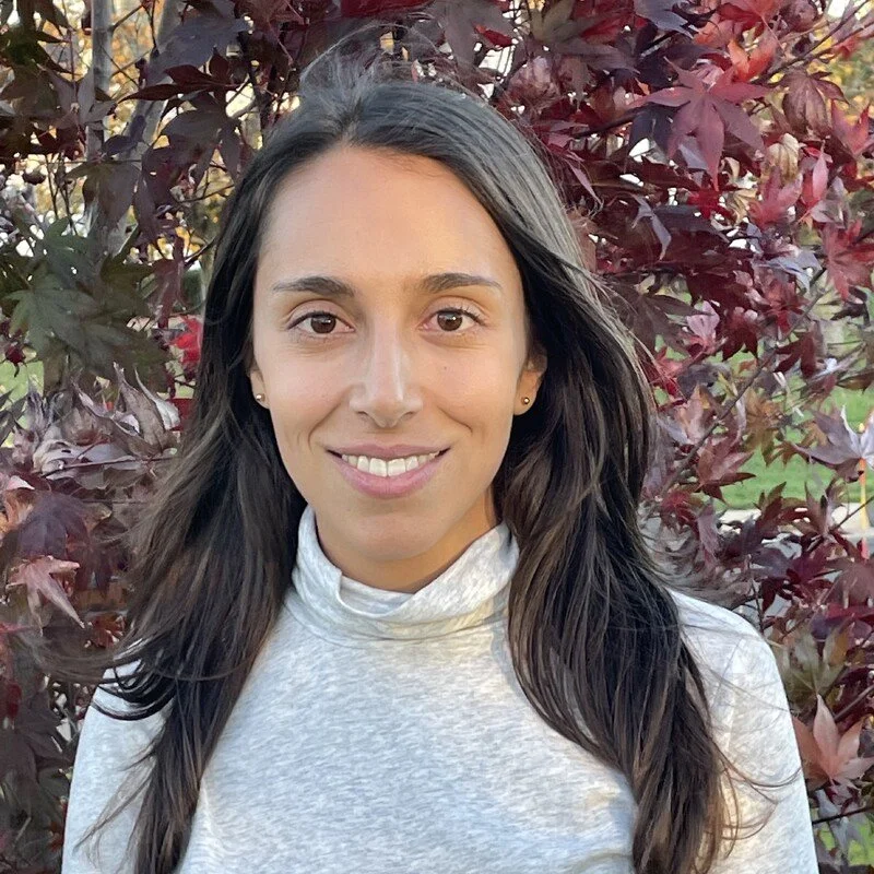 A young woman with long, dark hair smiling at the camera outdoors, standing in front of vibrant red and green leaves.