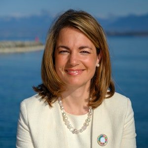 Smiling woman with shoulder-length light brown hair, wearing a white blazer and pearl necklace, standing outdoors near a body of water.