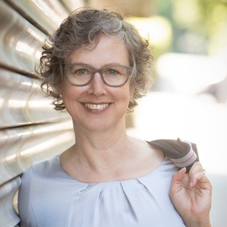 A smiling woman with curly short hair and glasses, wearing a light-colored top, holding a jacket over her shoulder in an outdoor setting with blurred background.