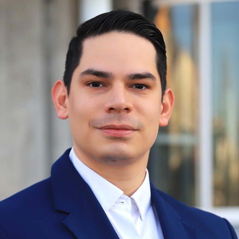 Headshot of a young man with black hair, wearing a navy blue suit and white shirt, standing outdoors in front of a building.
