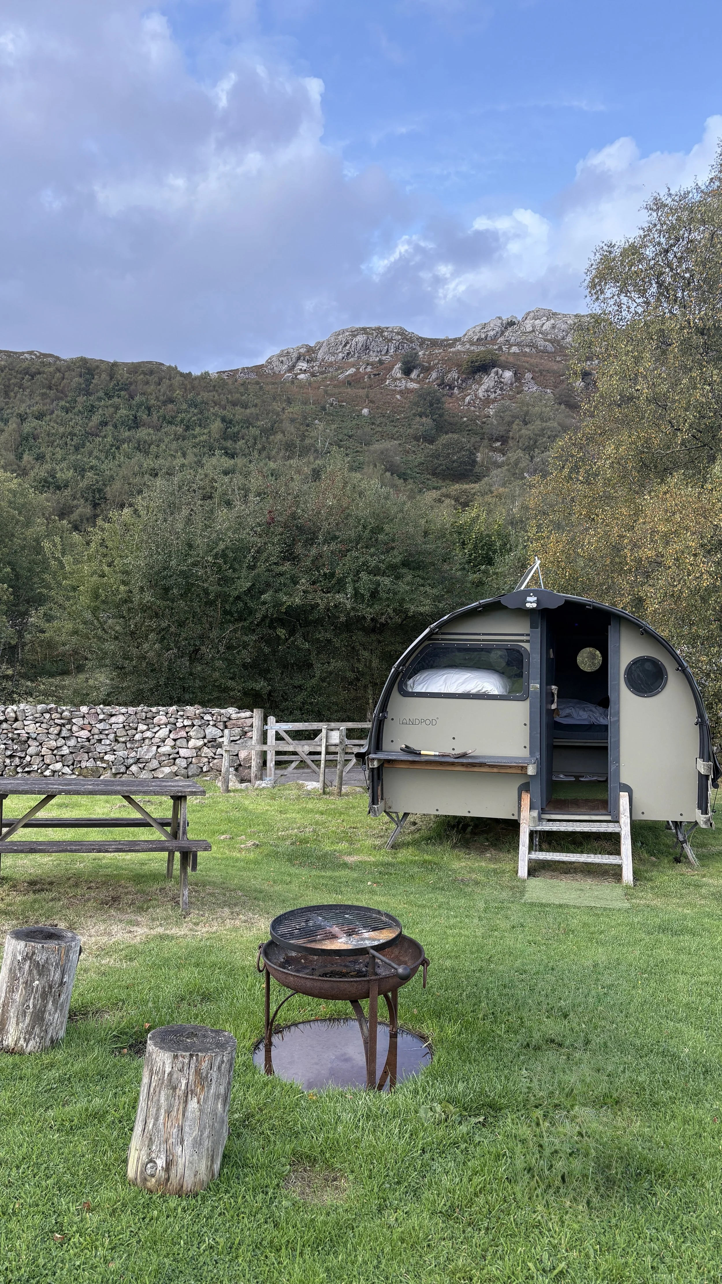 A small, rounded camper trailer parked on a grassy area with a picnic table and a fire pit in front. In the background, there are green trees and a rocky hillside under a partly cloudy sky.