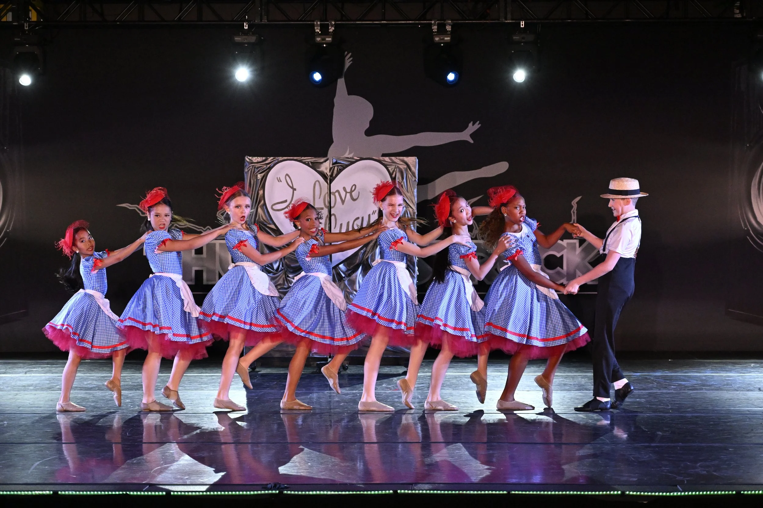 Group of young girls in blue checkered dresses with red accents dancing on stage, holding hands, with a girl in a straw hat at the right end.