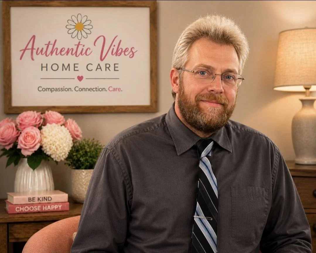 A middle-aged man with a beard, glasses, and blond hair, wearing a dark gray shirt and a striped tie, sitting in an office or therapy room. Behind him, a flowers arrangement, a framed sign reading 'Authentic Vibes Home Care' with a daisy, and a lamp are visible.