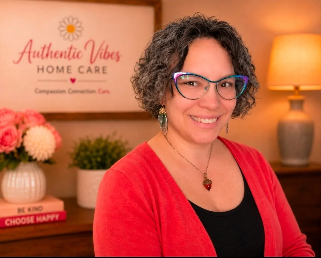 Kristi smiles at the camera. Her dark hair is short and curly. She wears colorful glasses, a necklace, and an orange cardigan.