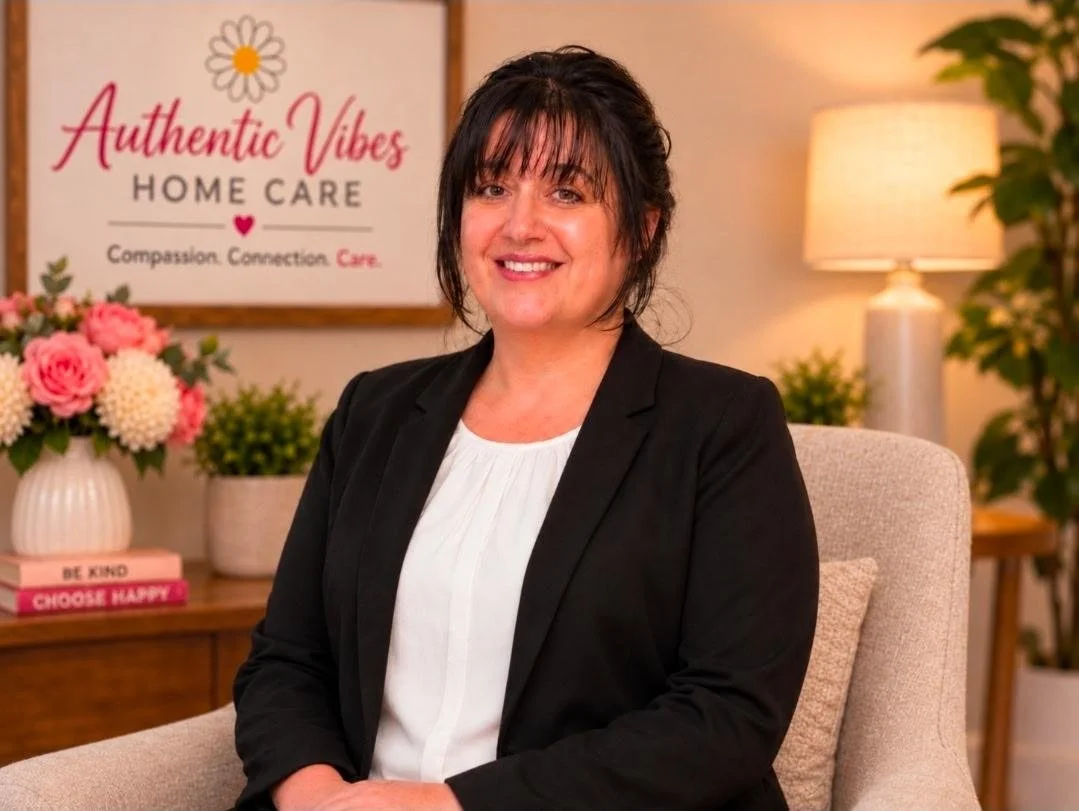 A smiling woman in a black blazer and white top sitting in a cozy home care setting. Behind her, a framed sign reads "Authentic Vibes Home Care" with the slogan "Compassion. Connection. Care.". There are decorative flowers and plants on the wooden table, and a small sign mentioning "Care, Comfort, Compassion.".