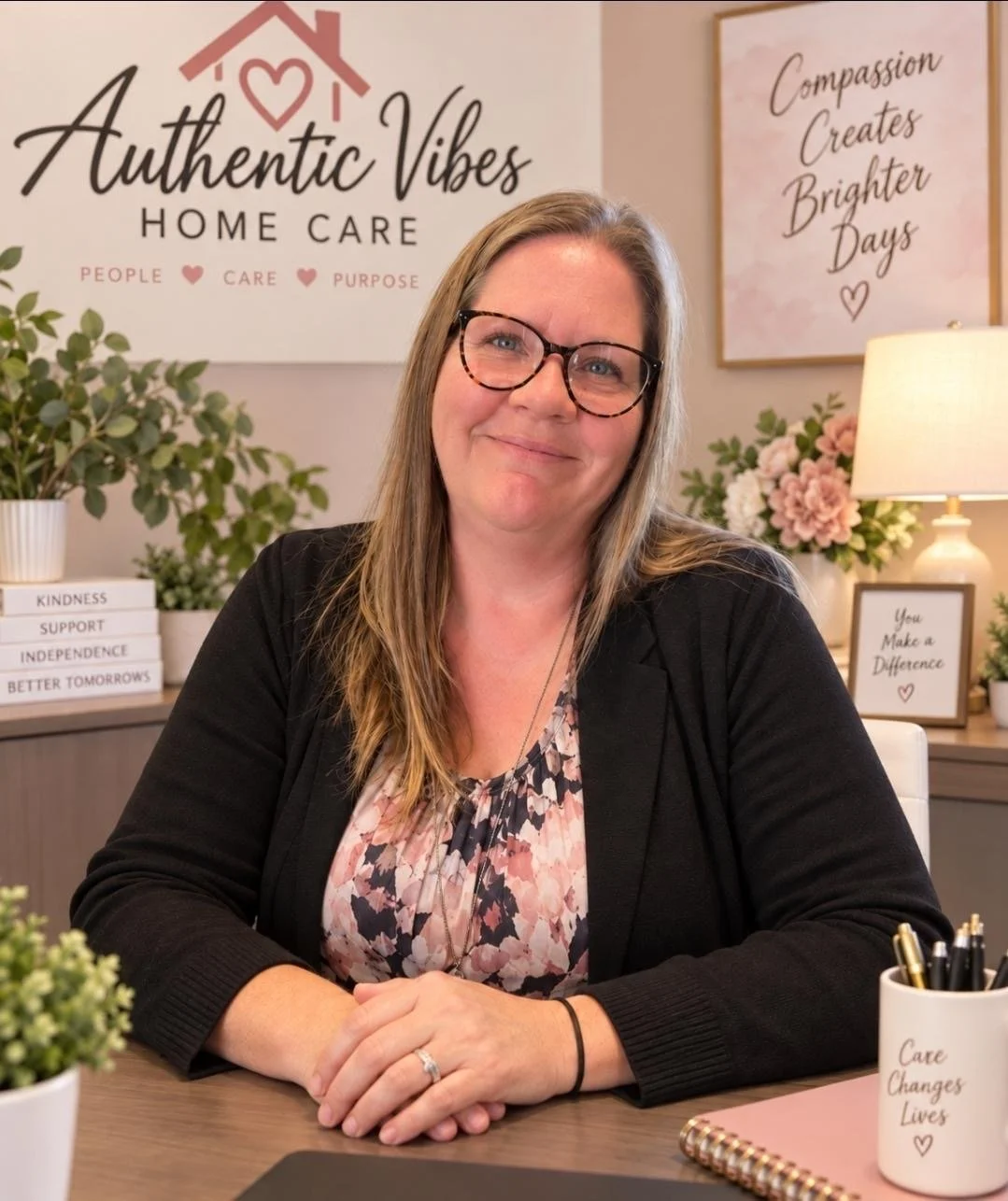 Rebecca smiles at the camera, hands folded and head tilted. Her long hair is loose and she wears a pink printed blouse and a black cardigan.