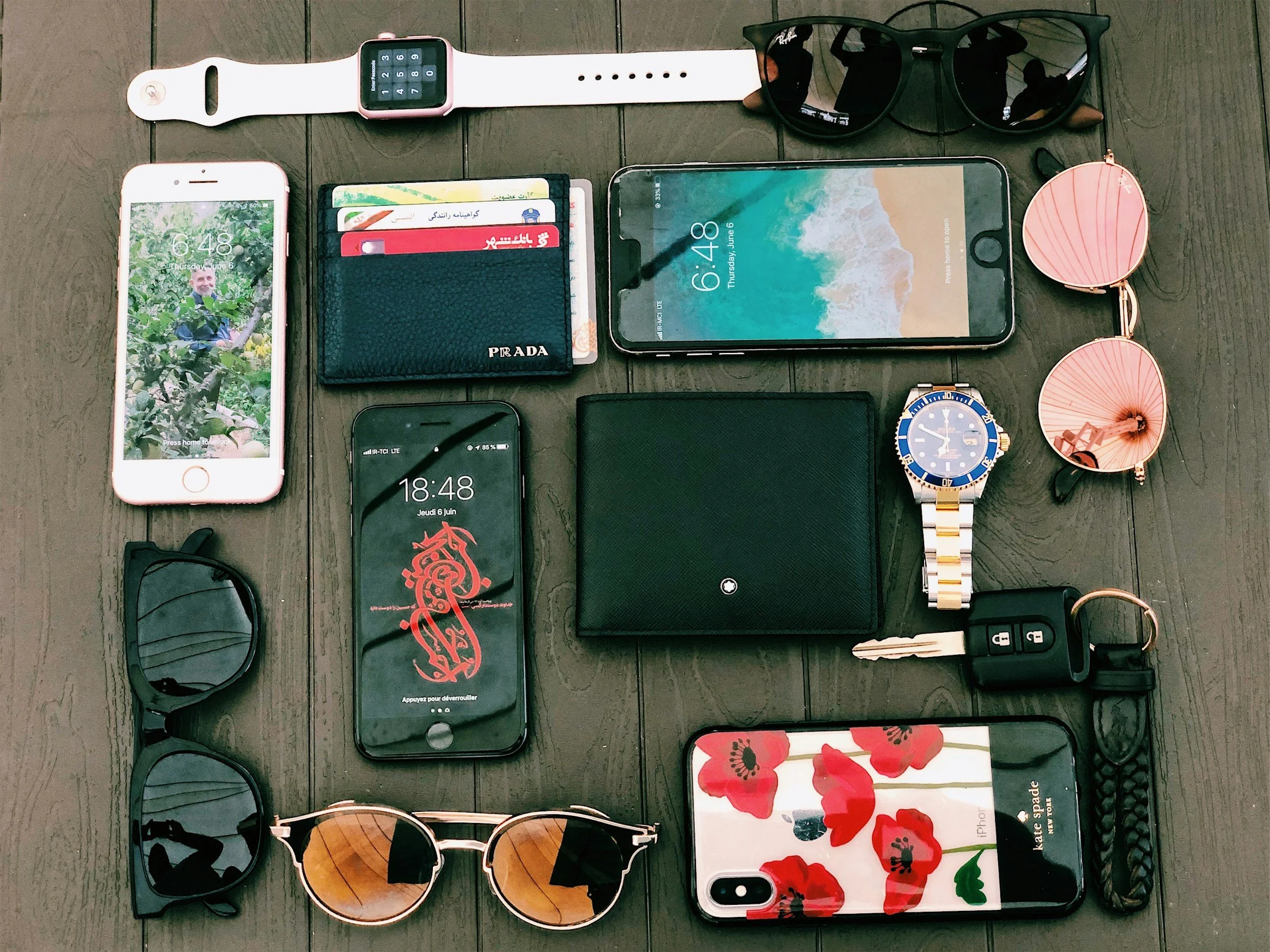 Collection of personal accessories and electronic devices on a wooden surface, including smartphones, sunglasses, a wristwatch, a wallet, a key, a water bottle, and miscellaneous small items.