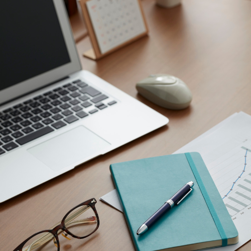 A workspace with a laptop, wireless mouse, glasses, a blue notebook, a pen, printed charts, and a framed calendar on a wooden desk.
