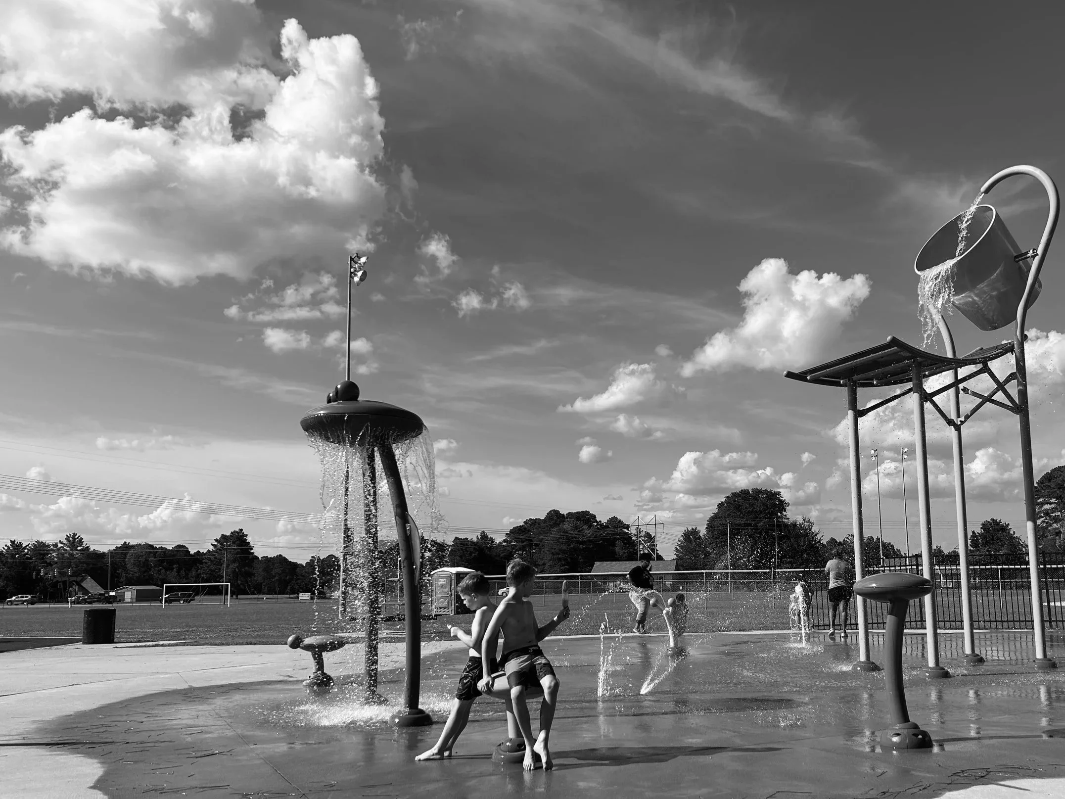 Children playing in splash park with water sprayers and water buckets, with cloudy sky and sports fields in the background.