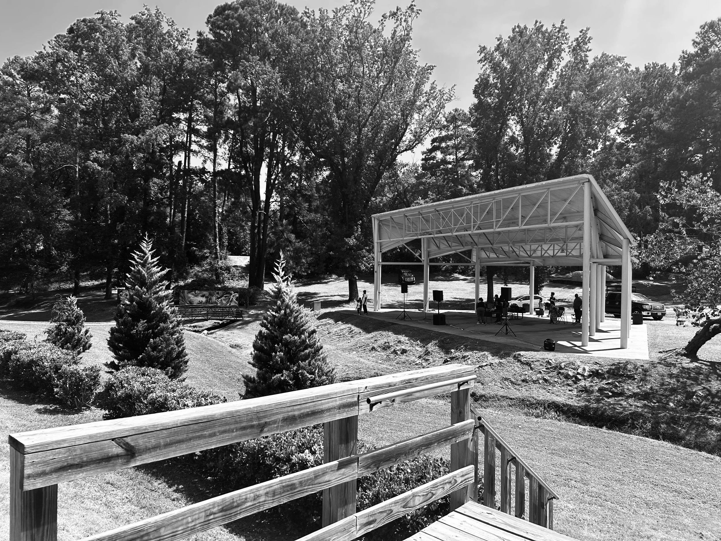 Black and white photo of an outdoor concert area with a covered stage and musical instruments, surrounded by trees and greenery.