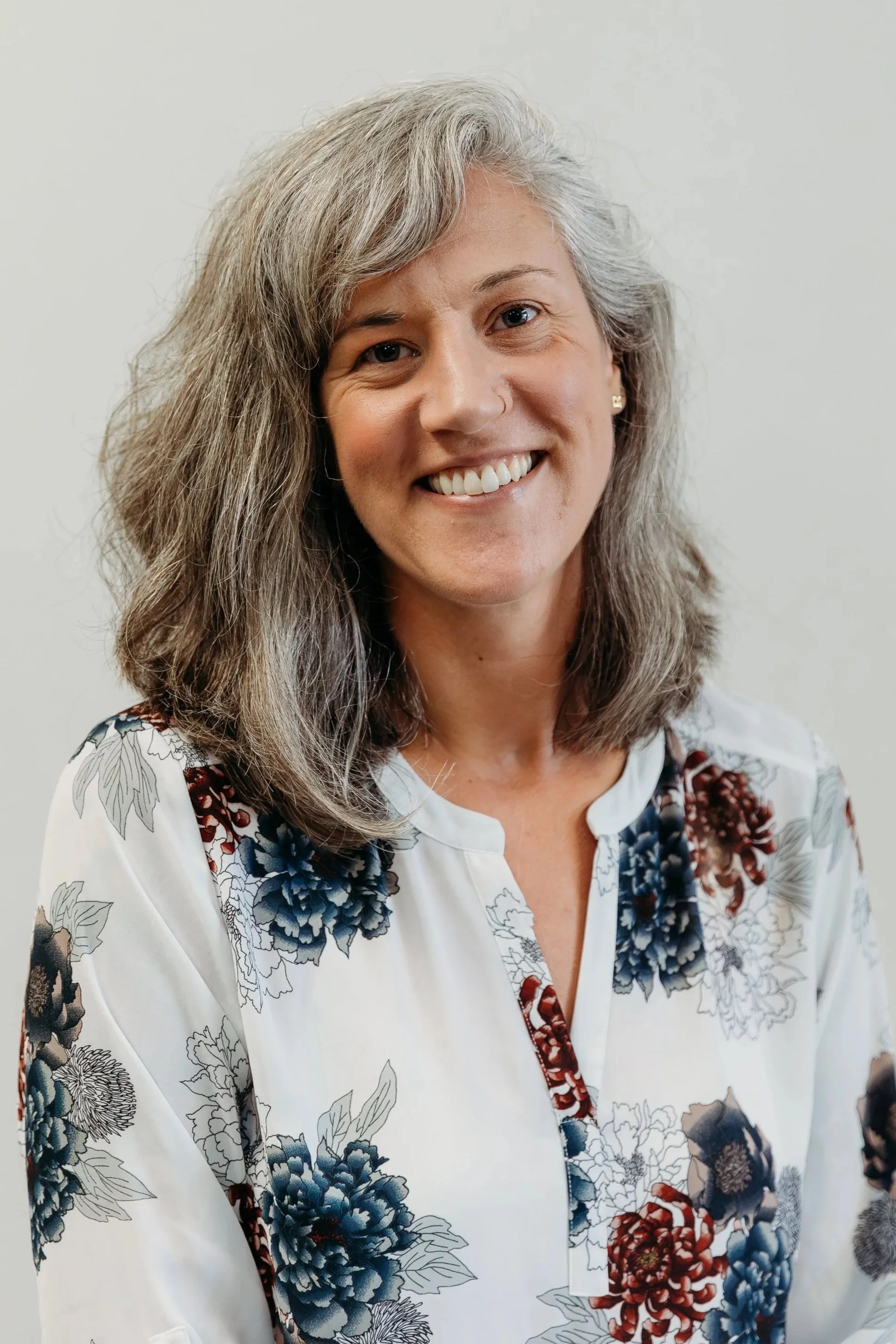 A woman with gray hair and a nose ring smiling, wearing a white floral blouse with blue, red, and black flowers.